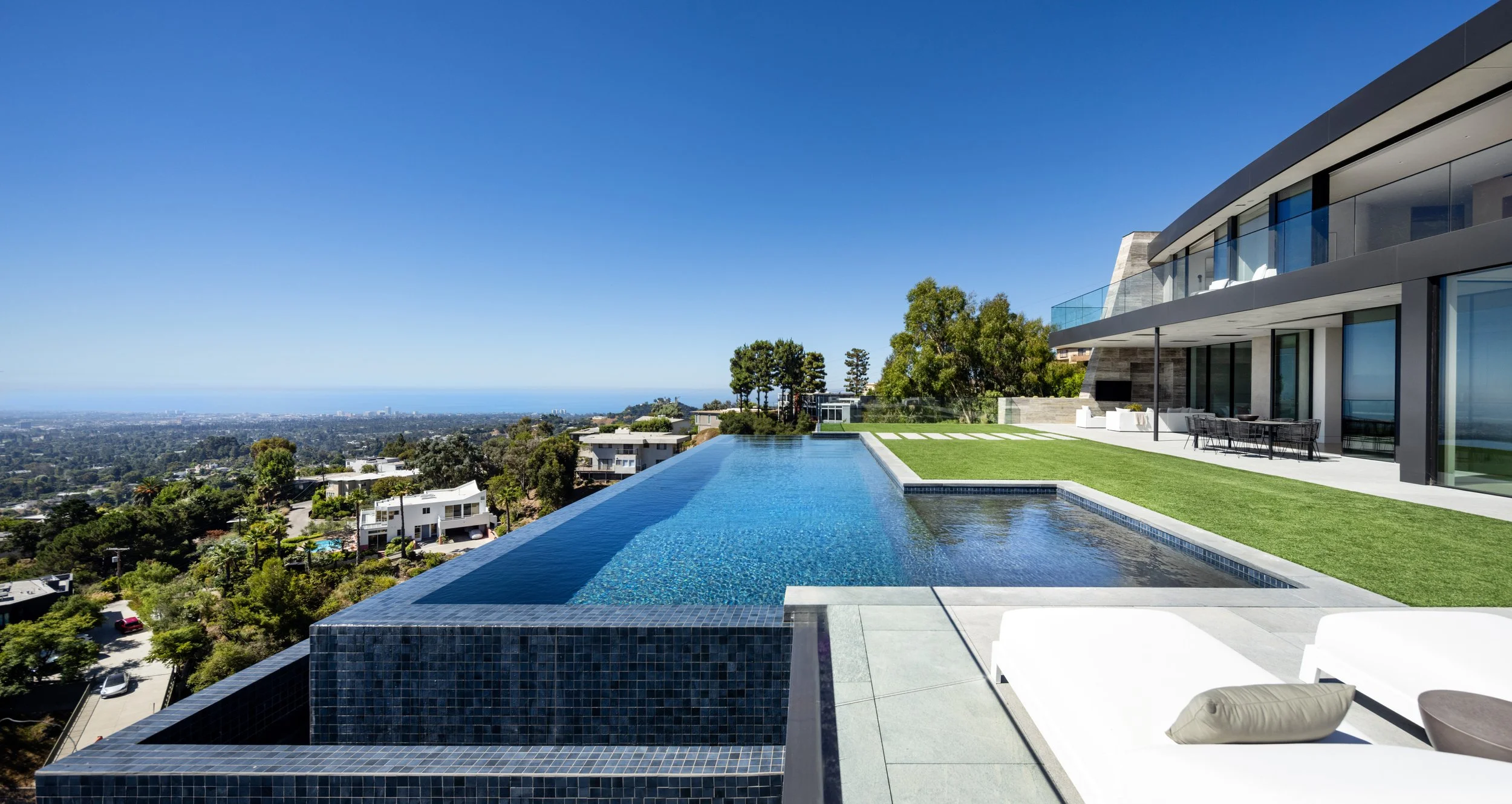 Luxury modern house with a rooftop infinity pool, outdoor seating, and panoramic city view under clear blue sky.