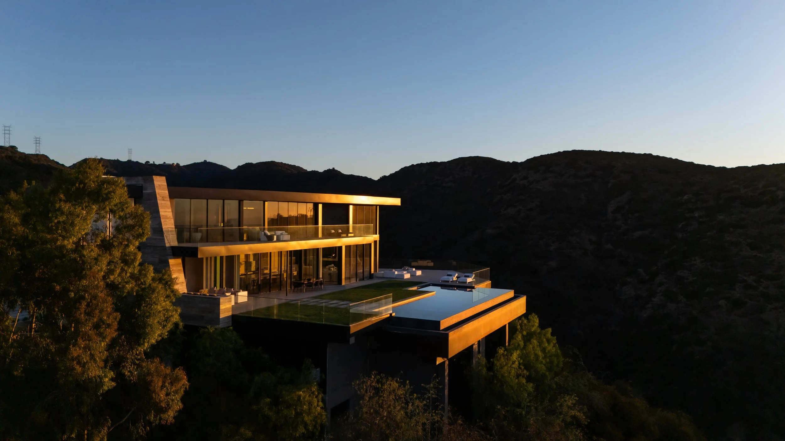 Modern house with large glass windows and an outdoor pool situated on a hillside at sunset, with hills in the background and trees in the foreground.