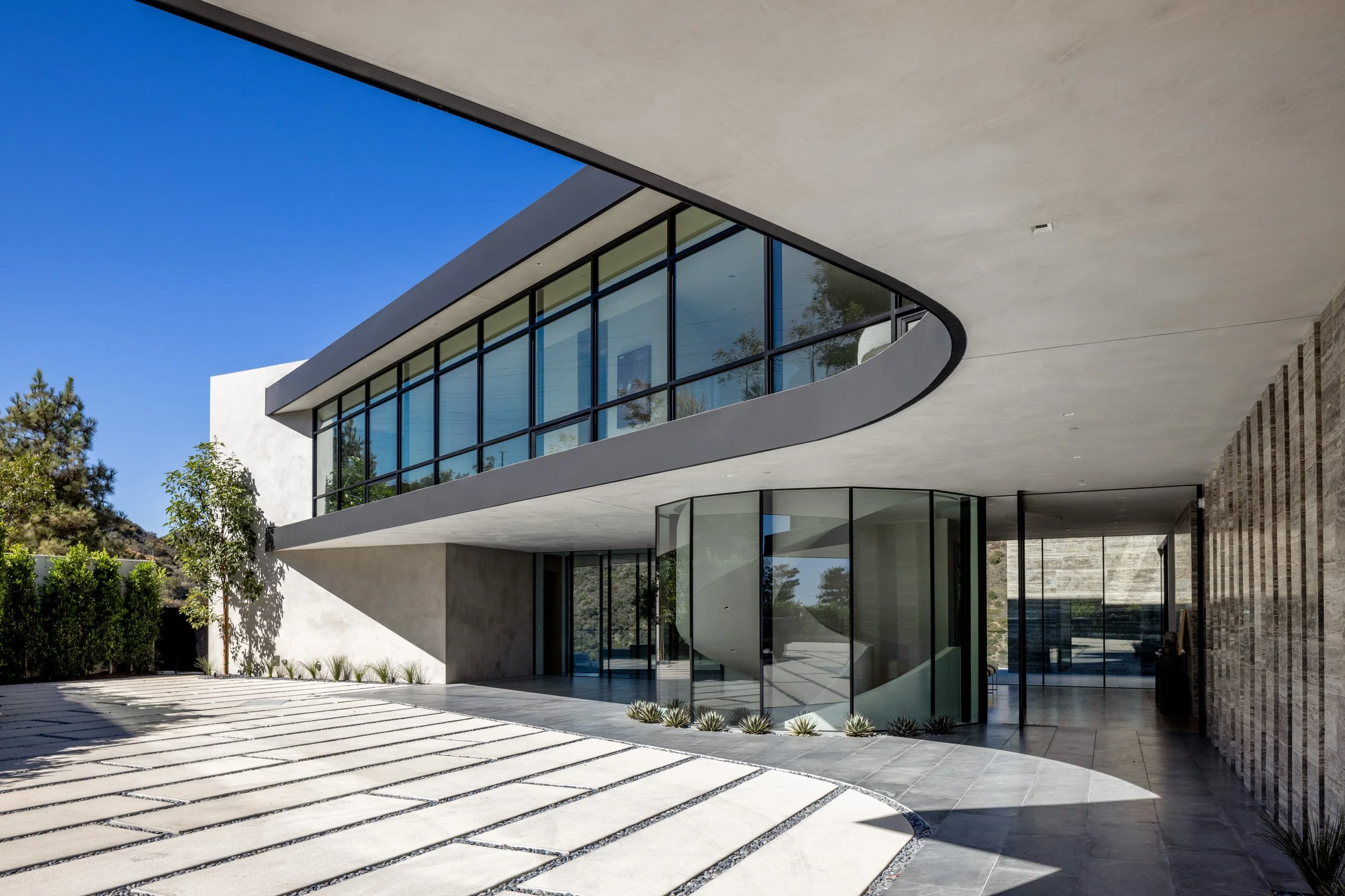 Modern house with curved architectural design, large glass windows, and outdoor landscaping, featuring a paved courtyard and minimalist landscaping.