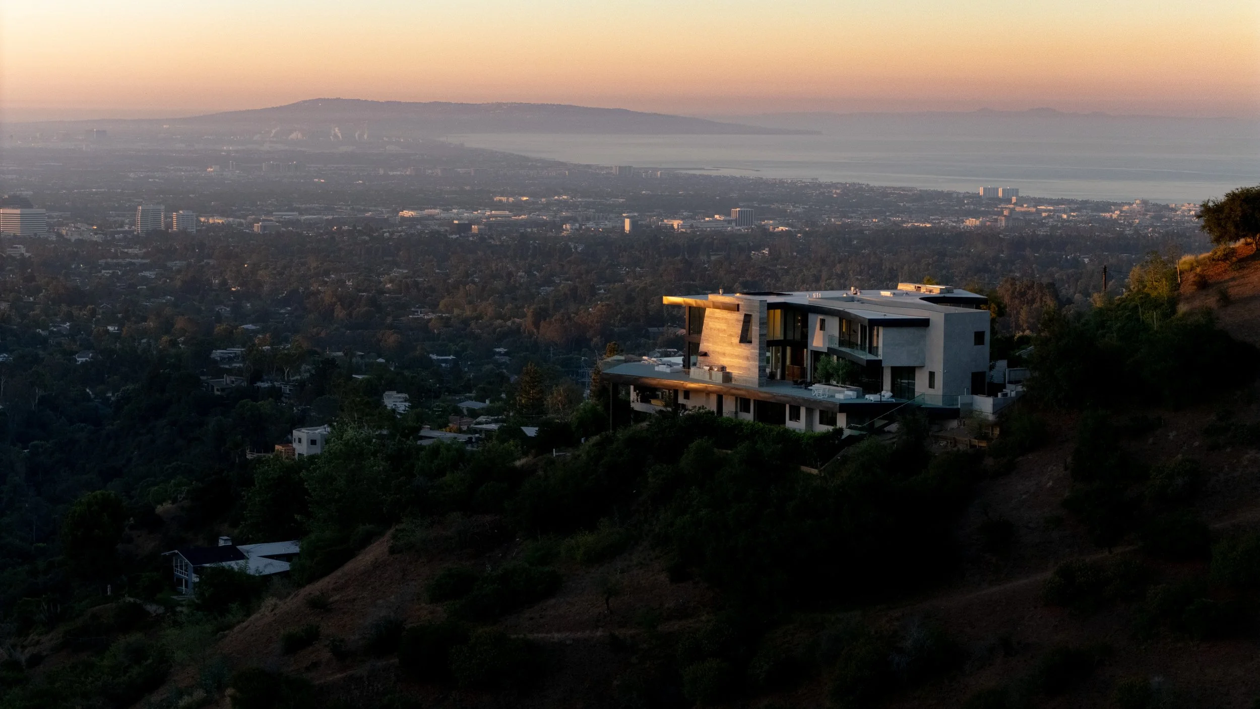 Modern house on hillside overlooking a city and water with mountains in the background at sunset.