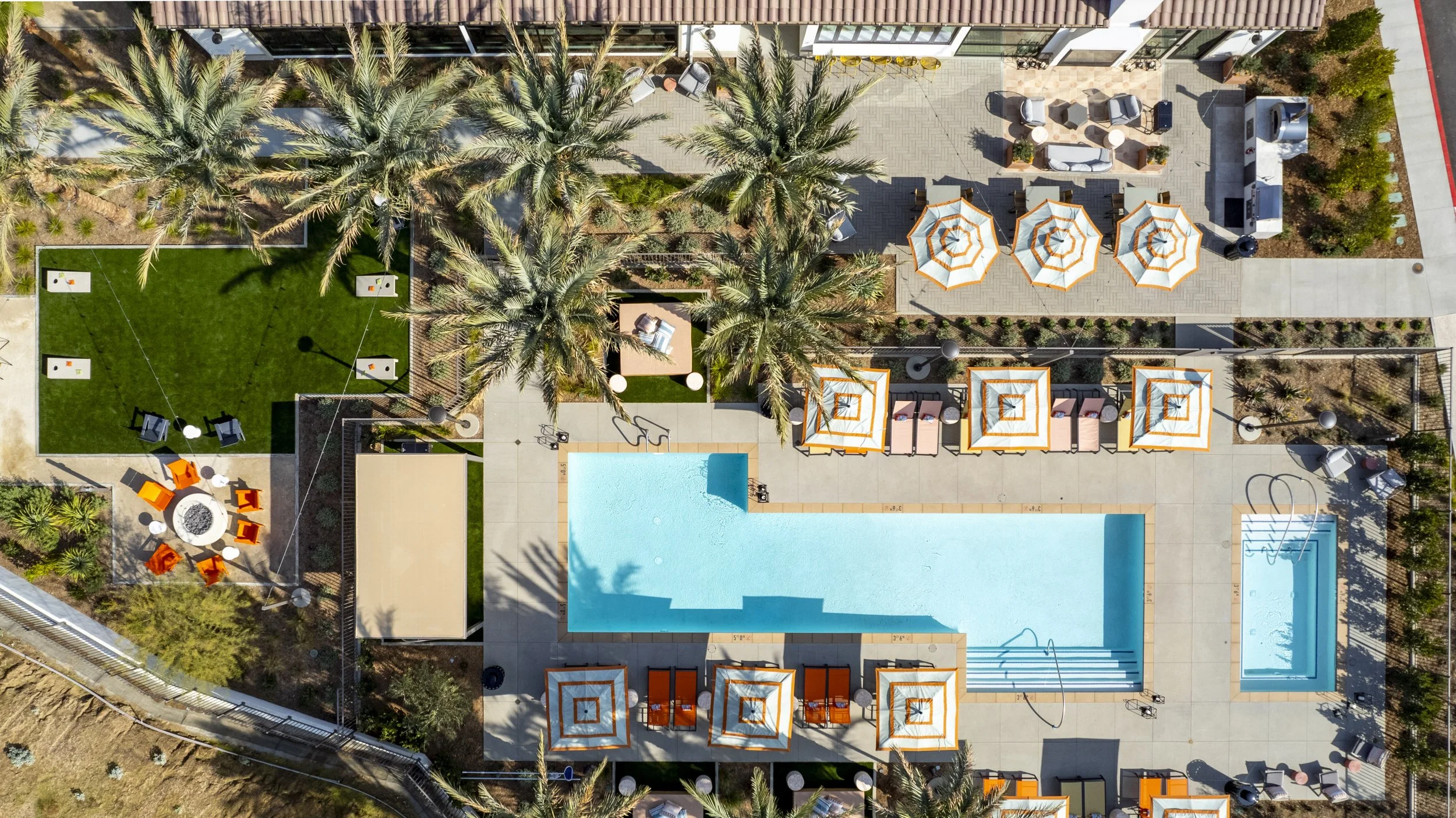 Aerial view of a swimming pool area with lounge chairs, umbrellas, and surrounding greenery, including palm trees and a small lawn with outdoor seating, in a landscaped backyard or resort setting.