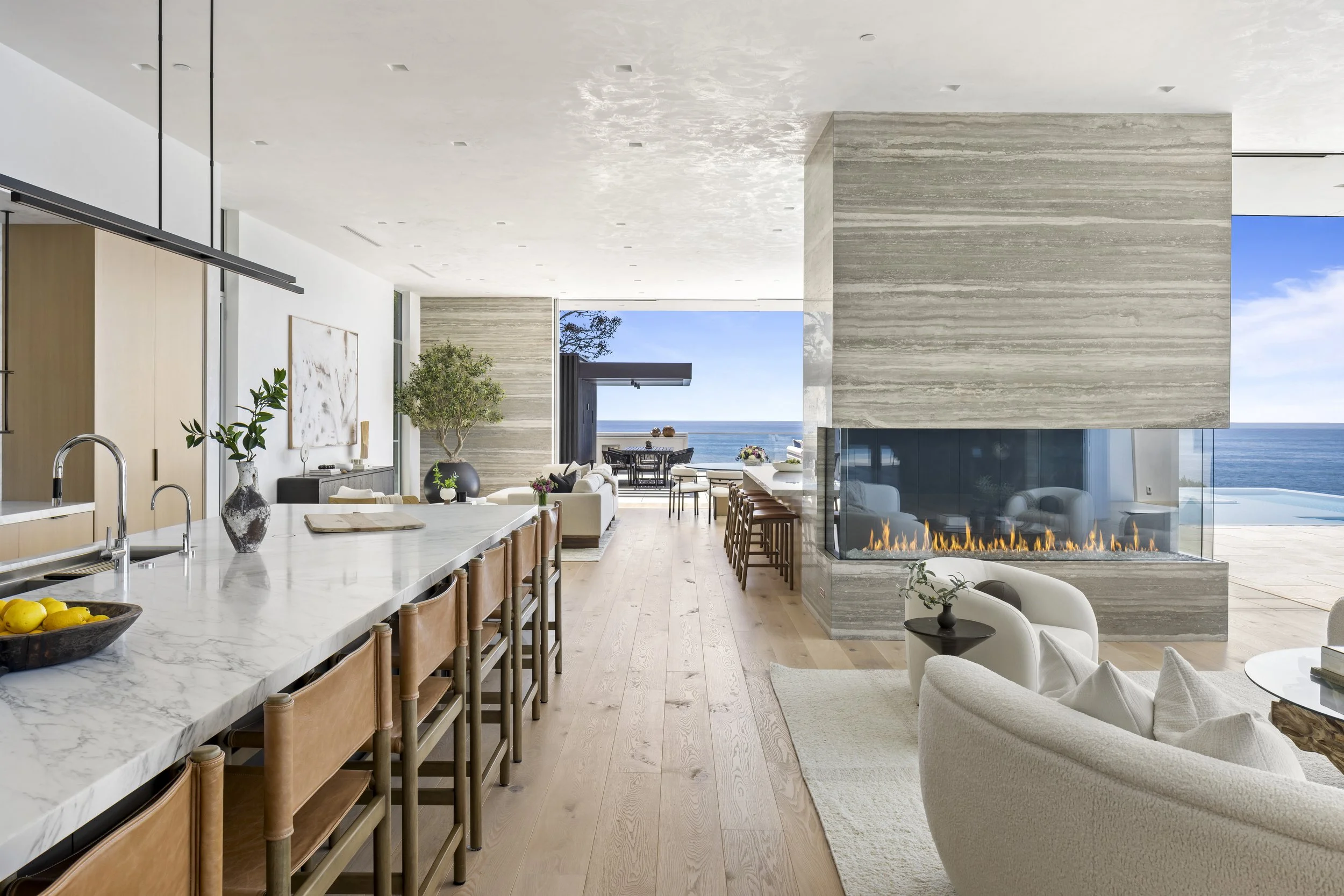 Open-concept living and kitchen area with a view of the ocean, featuring a modern fireplace, marble kitchen island with wooden barstools, and minimalistic furniture with decorative plants.