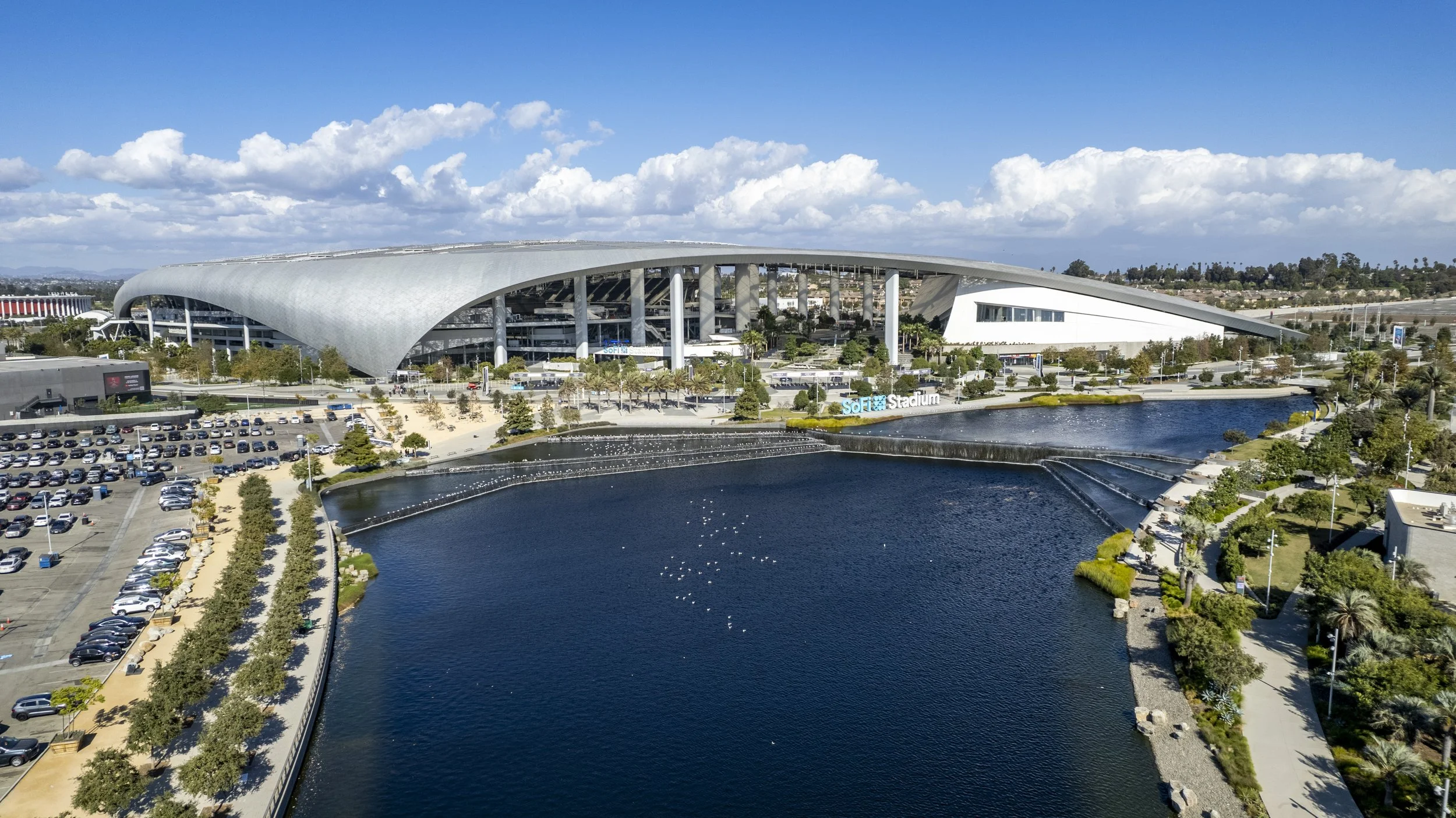 Aerial view of SoFi Stadium with water feature and parking lot in the foreground, clear blue sky with clouds in the background.