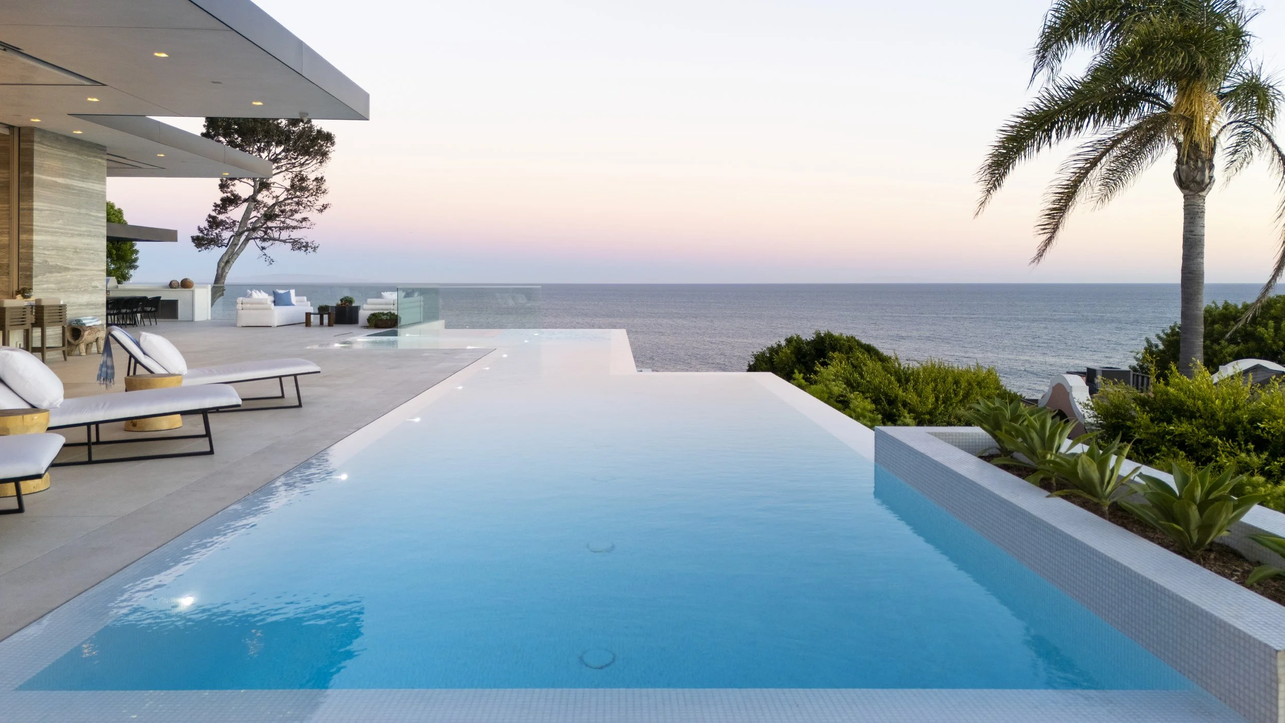 Luxury infinity pool overlooking the ocean at sunset, with lounge chairs, lush green plants, a palm tree, and modern patio furniture.