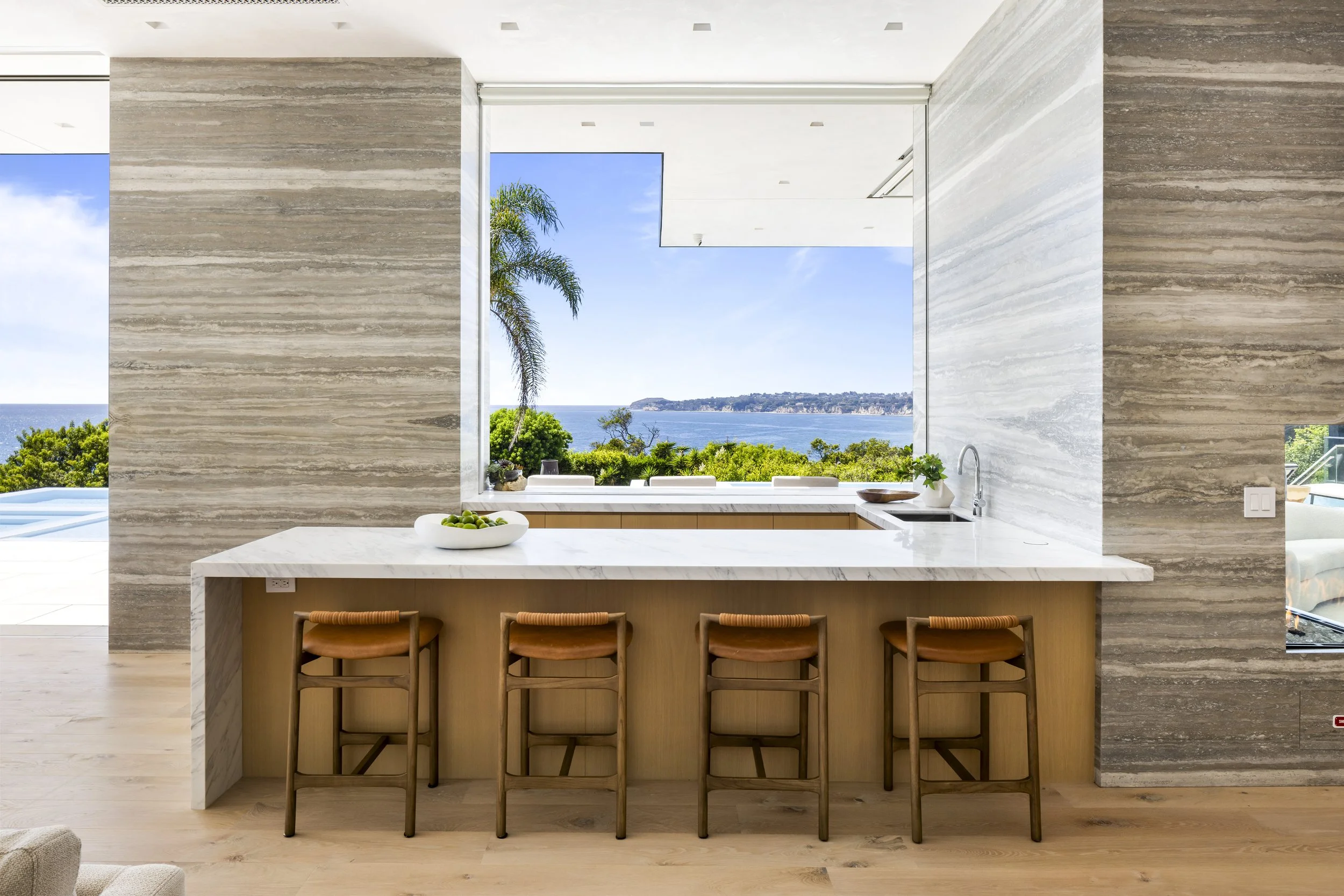 Modern kitchen with marble countertops and bar stools overlooking a scenic ocean view through a large open window.