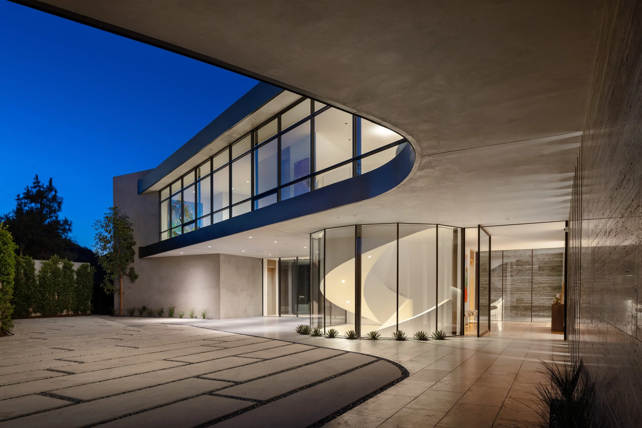Modern house at night with large glass windows, a spiral staircase inside, and a spacious exterior patio.