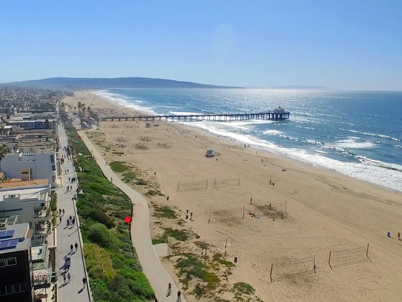 A sunny beach with a pier extending into the ocean, with people walking along the shore and on nearby pathways, and beachfront buildings lining the coast.