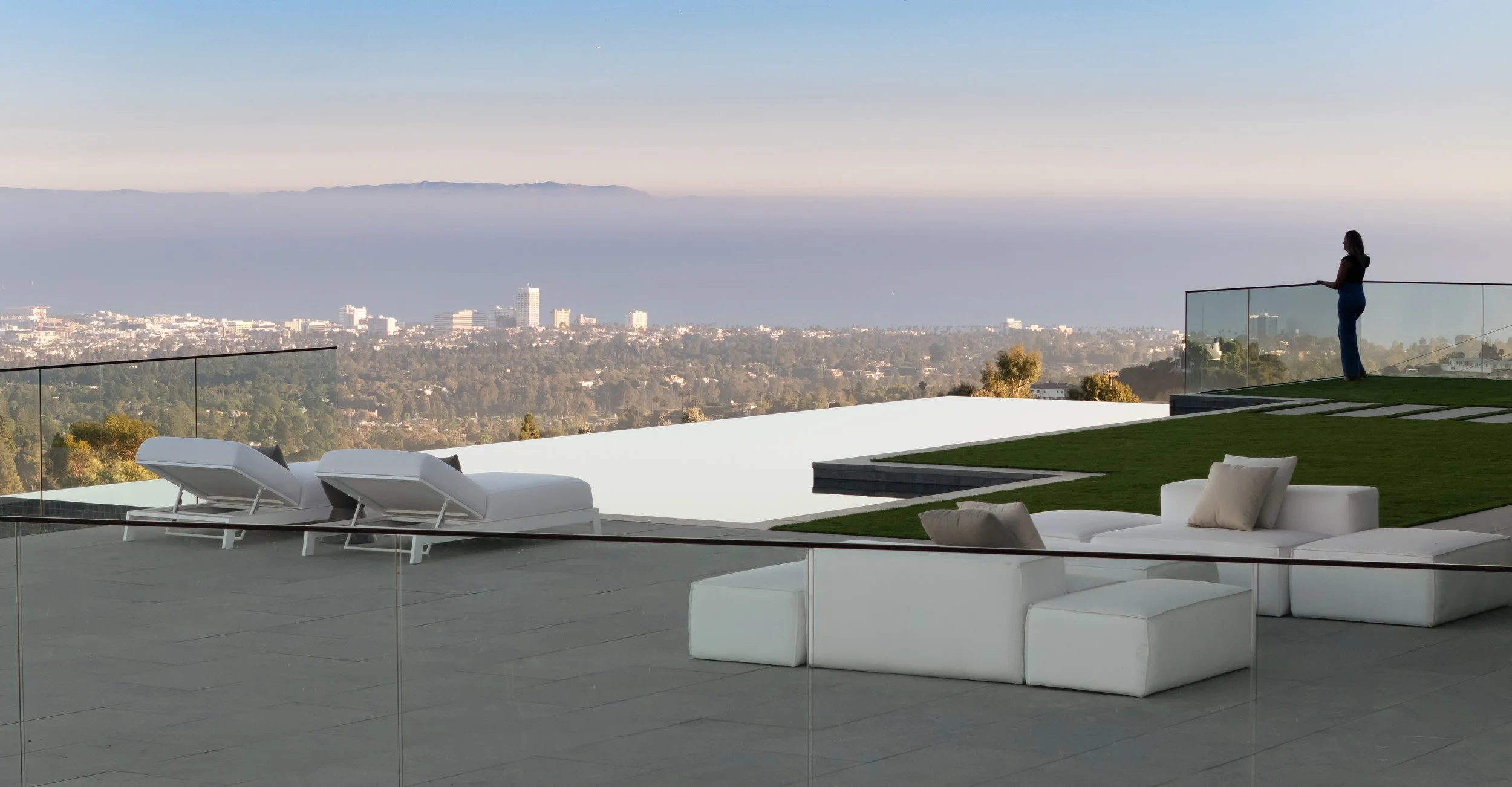 A modern outdoor terrace overlooking a cityscape with a woman standing near a glass railing, with a view of mountains and buildings in the distance. There are white lounge chairs and a white sectional sofa with pillows on the terrace.