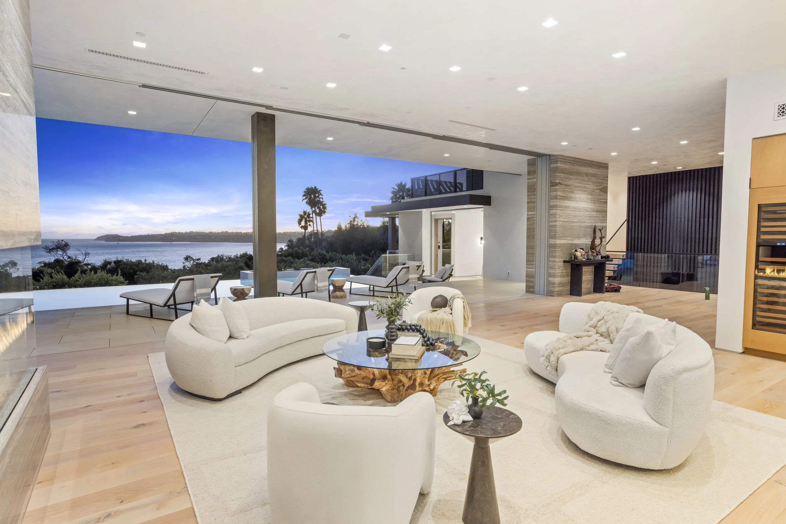 Modern living room with white plush seating, a wooden centerpiece table, and a view of an outdoor patio and water at sunset.