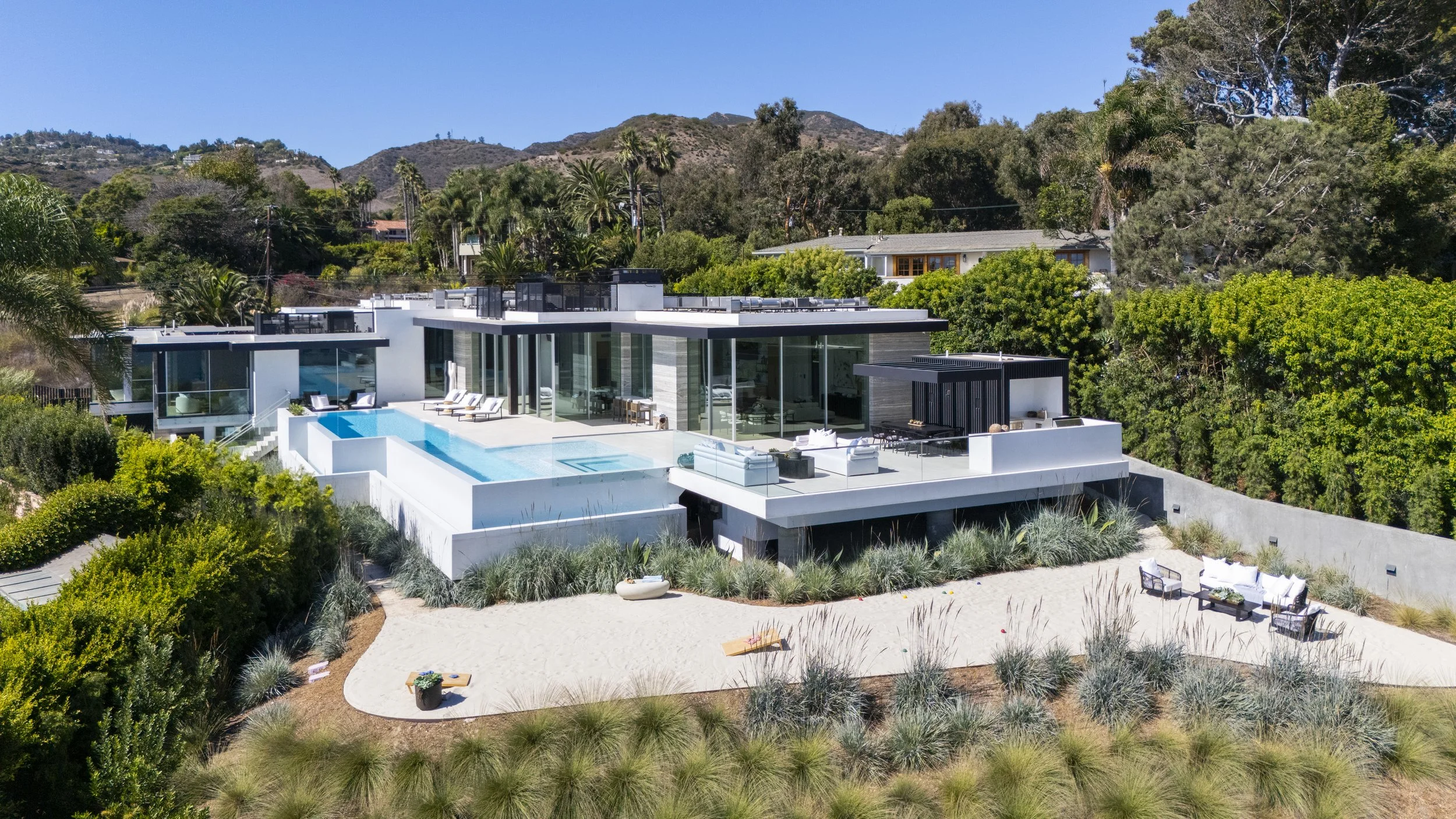 Modern house with flat roof, large glass windows, outdoor pool, and patio furniture, located on a hillside with lush greenery and trees in the background under a clear blue sky.