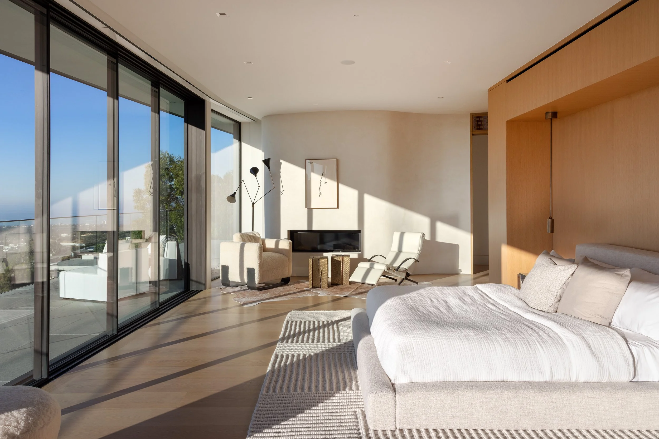 Modern living room with large glass sliding doors, natural sunlight, a bed, lounge chairs near the window, and minimalist decor.
