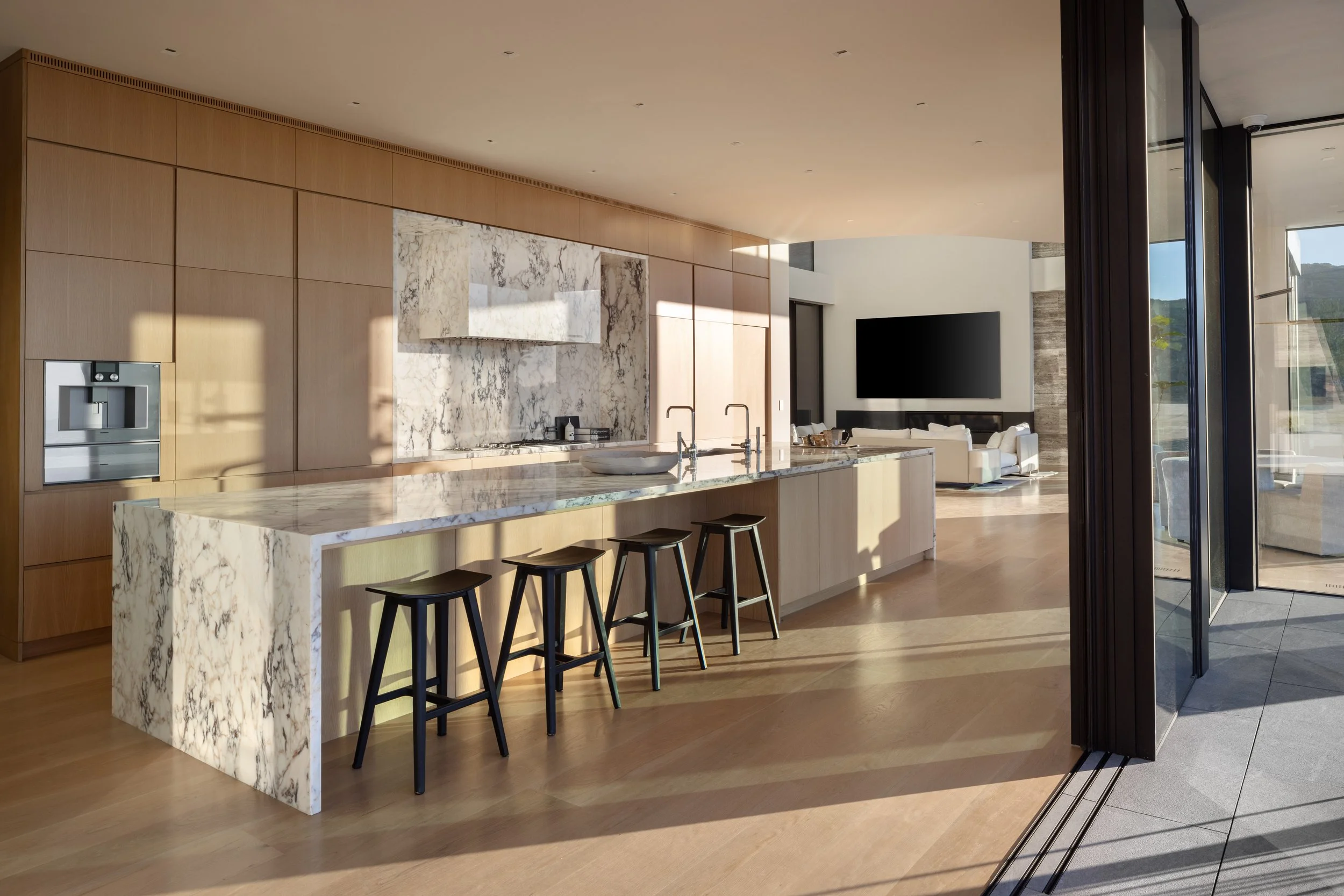 Modern open-concept kitchen with marble island and wood cabinets, leading to a living area with a large flat-screen TV and white sofa, illuminated by natural sunlight