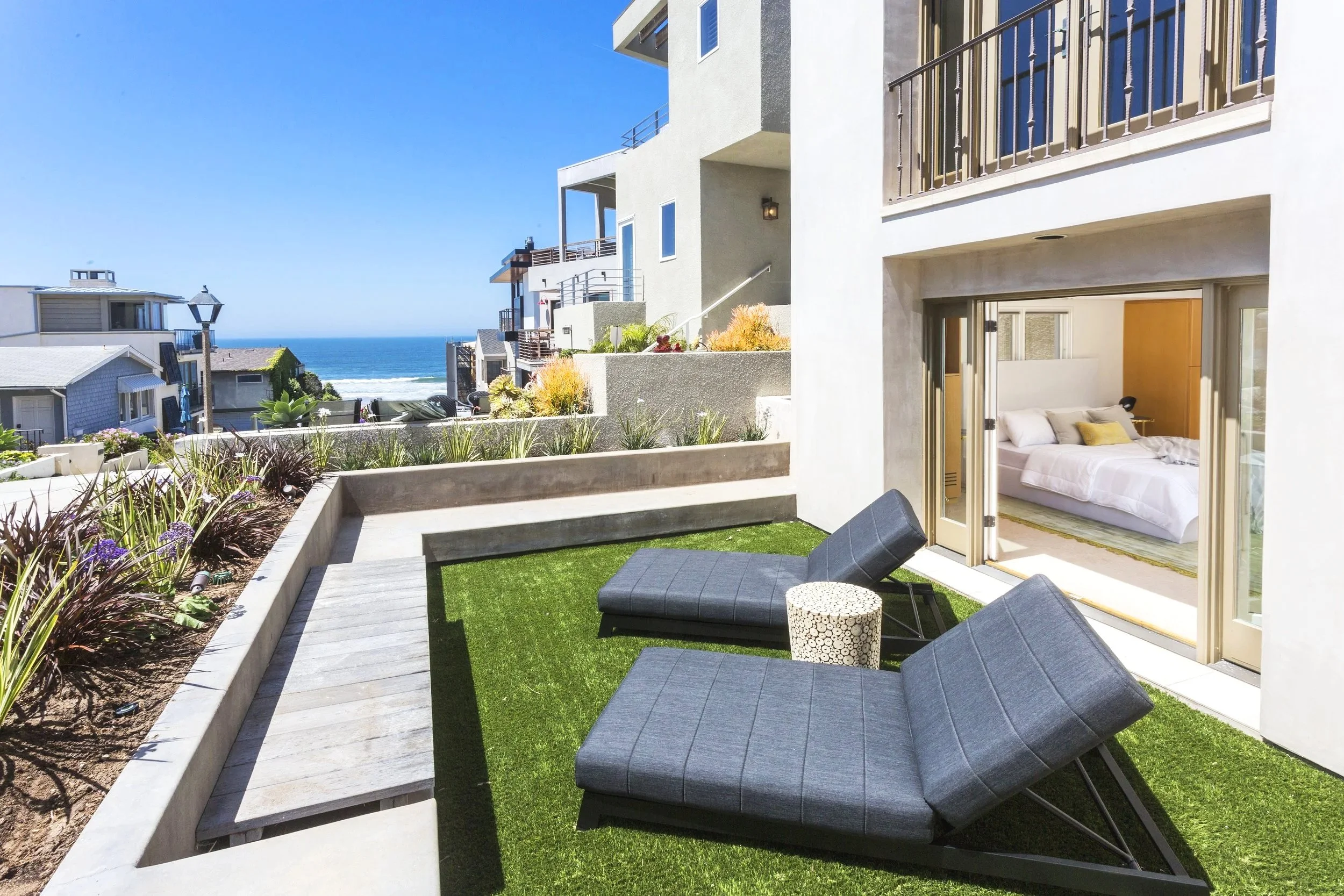 Beachside balcony with two black lounge chairs, small round side table, and view of ocean, neighboring houses, and blue sky.