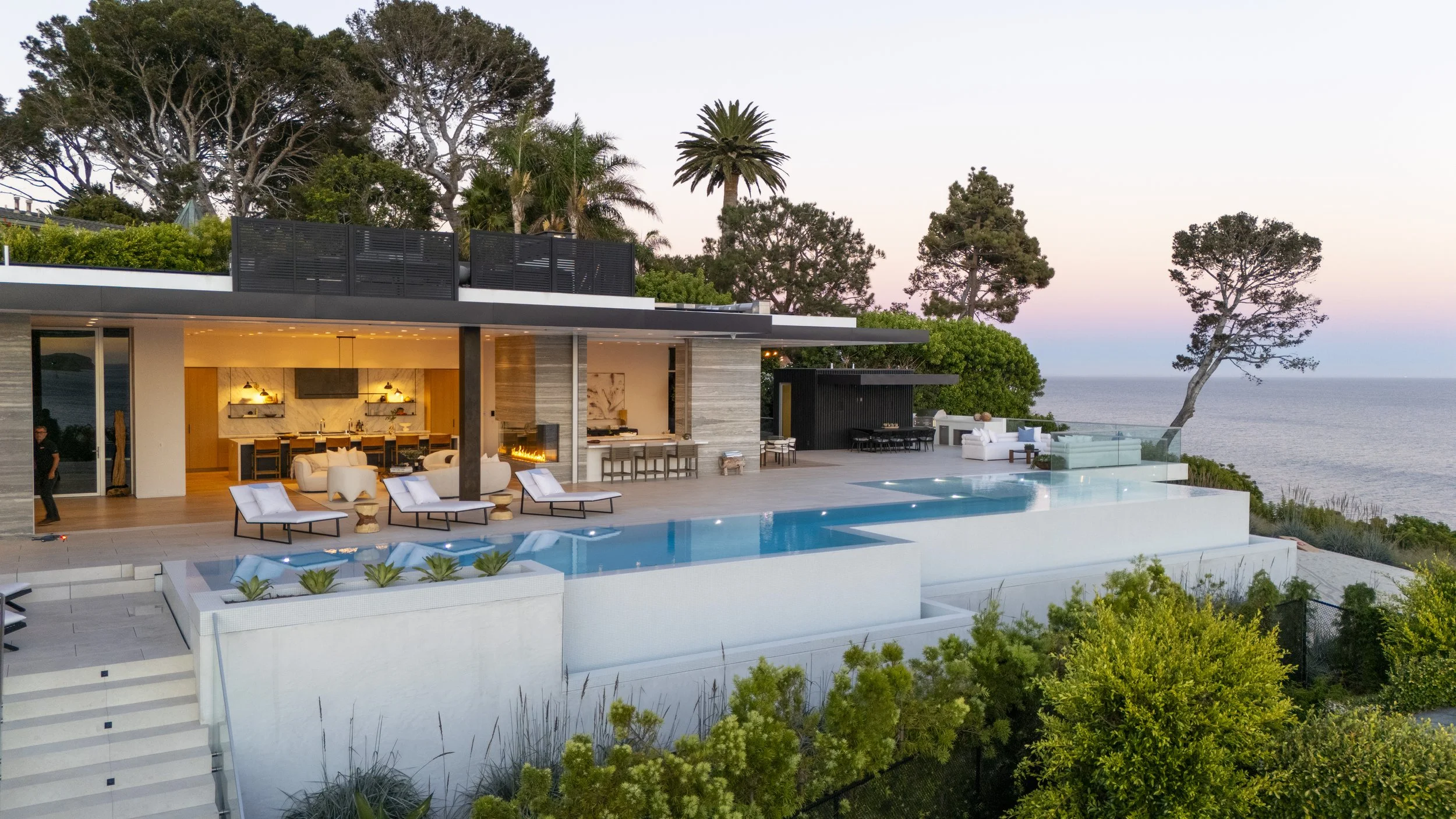 Modern house with an outdoor pool on a hillside overlooking the ocean, with a patio area, lounge chairs, and outdoor seating, surrounded by trees and greenery at sunset.
