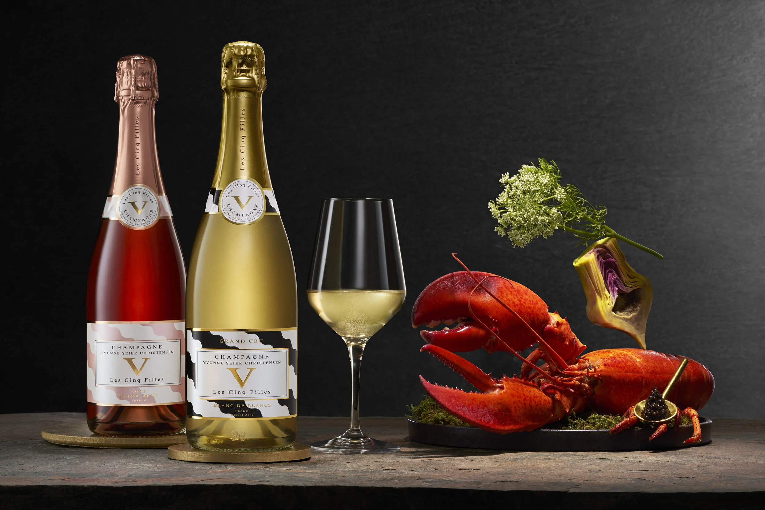  Photo of Les Cinq Filles Champagne bottle and glass with seafood arranges as a sculpture all sitting on a stone foreground.  