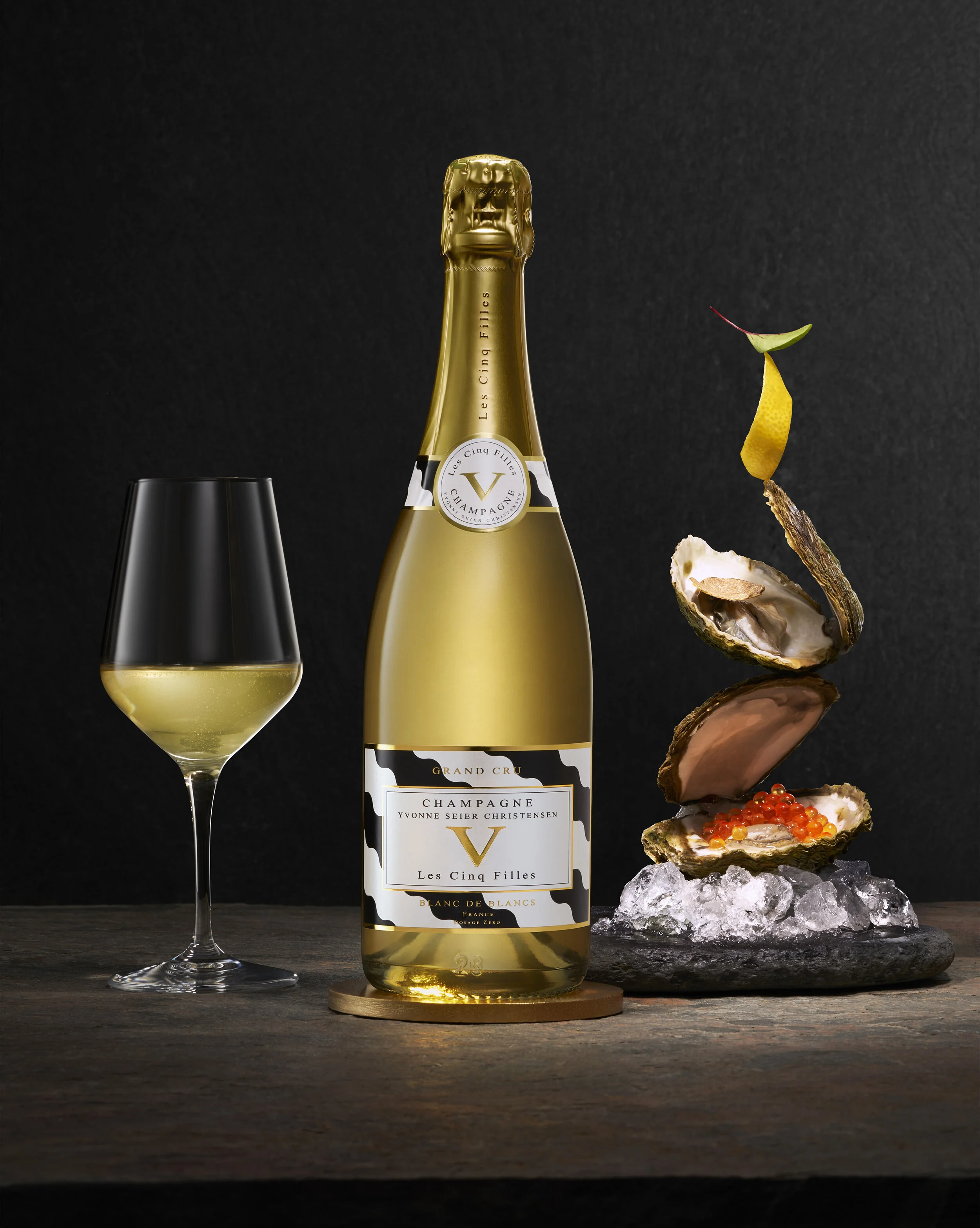  Photo of Les Cinq Filles Champagne bottle and glass with seafood arranges as a sculpture all sitting on a stone foreground.  