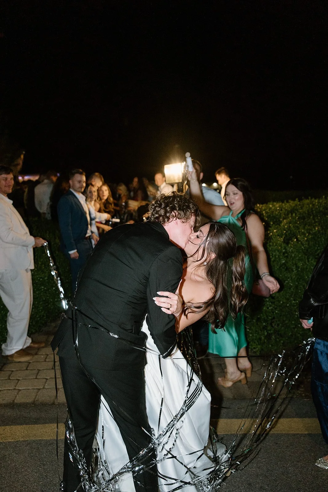 A newlywed couple sharing a kiss at night with guests surrounding them at a celebration, with some holding a phone and decorations.
