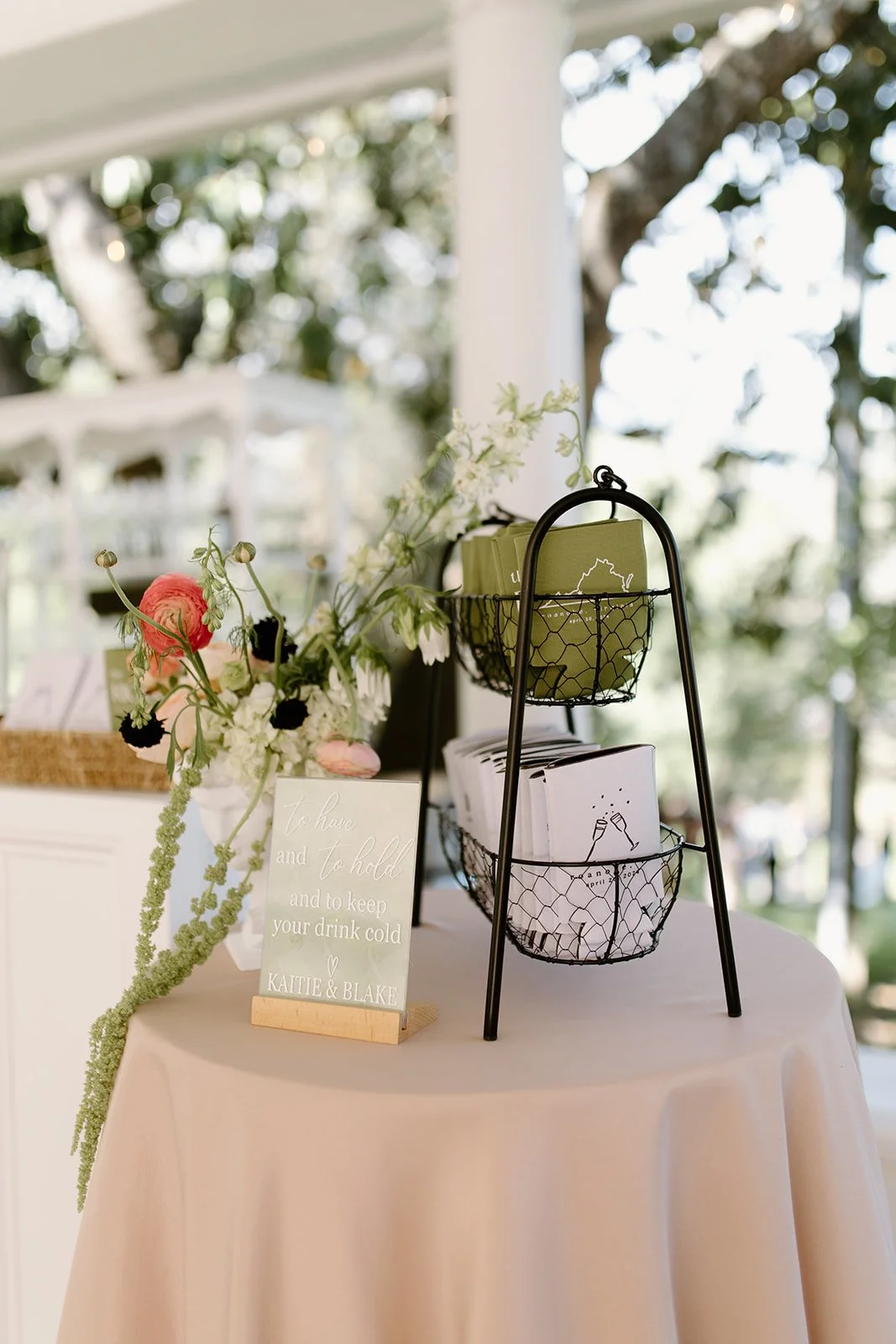 Decorative table with a small floral arrangement, a sign that says 'To have and to hold and to keep your drink cold, Katie & Blake,' and a two-tiered black metal stand holding green and white boxed items at an outdoor event.