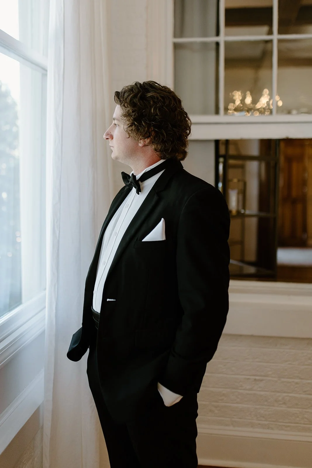 A man dressed in a black tuxedo with a white shirt and black bow tie, standing indoors near a window, looking outside.