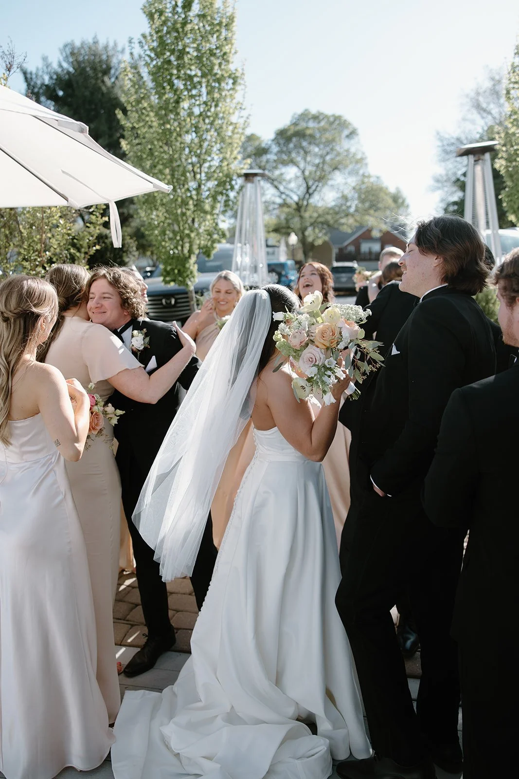 A bride in a white wedding dress holding a large bouquet of flowers, surrounded by smiling wedding guests outdoors on a sunny day.
