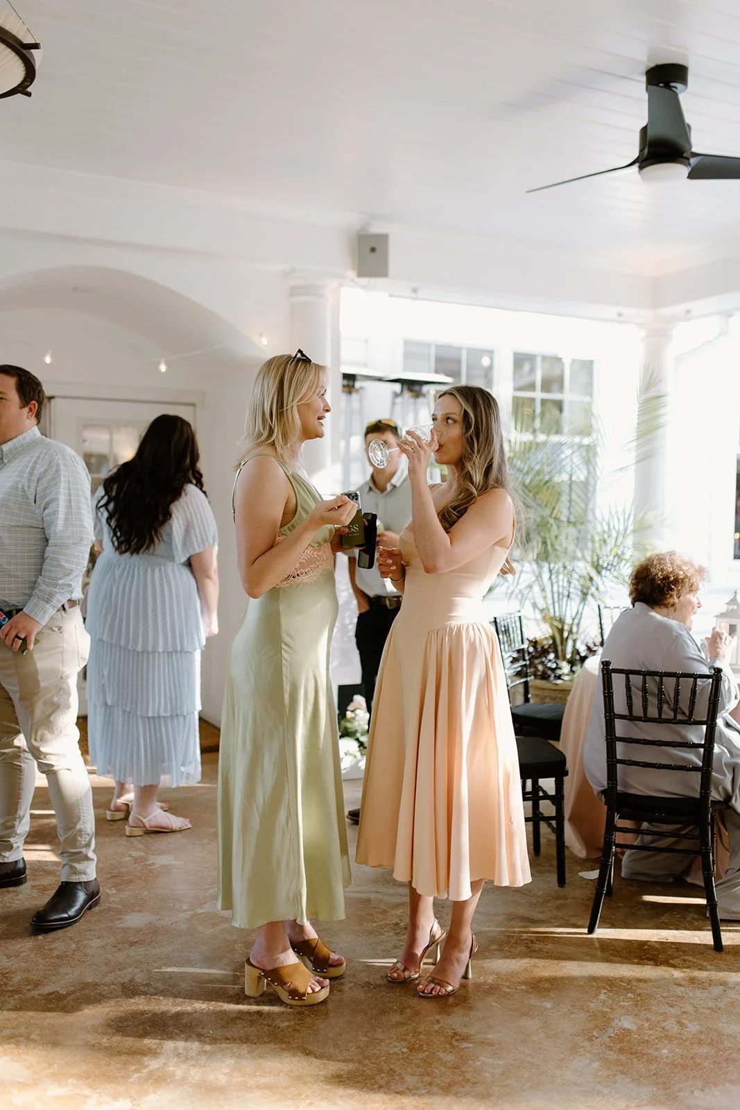 Two women in pastel-colored dresses are standing and chatting at a lively indoor event, with one drinking from a glass. Other guests are mingling in the background.