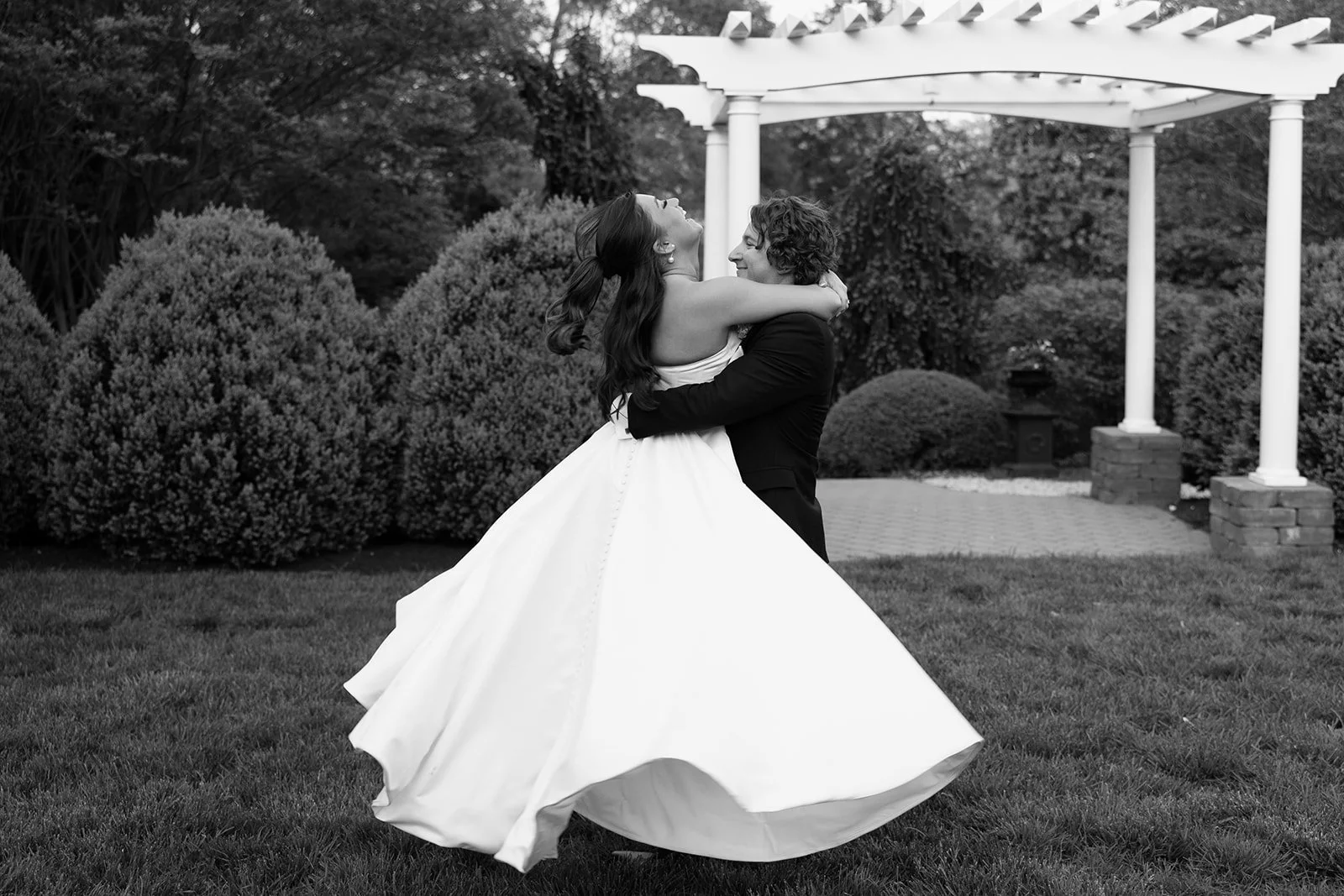 A bride and groom dancing outdoors in a garden, with the bride in a flowing white gown and the groom in a dark suit, surrounded by bushes and trees.