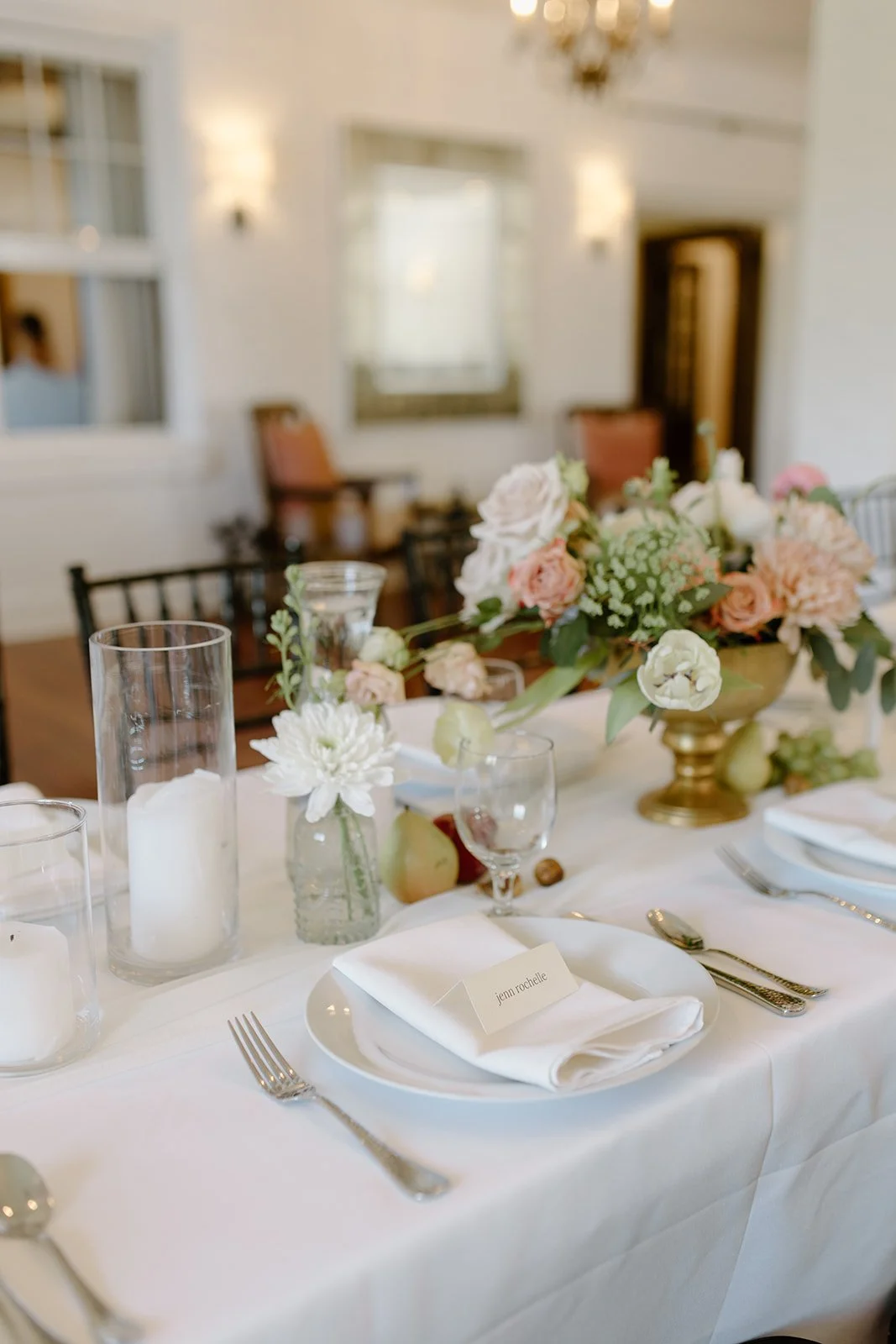 Elegant table setting with white tablecloth, floral centerpiece, candles, and place card reading 'Jenn Rochelle' in a decorated reception hall.