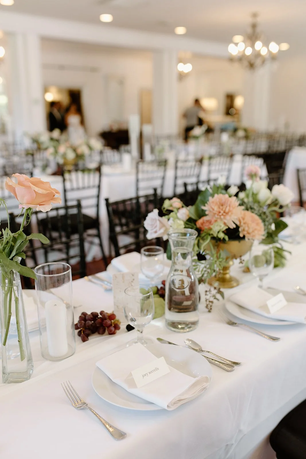 Elegant banquet table set with white tablecloths, floral centerpieces, plates with napkins and name cards, glasses, and silverware, in a decorated room with chandeliers.