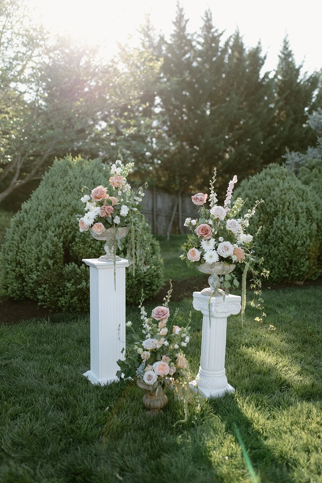 Three floral arrangements on white and beige pedestals outdoors in a garden with green bushes and trees, sunlight shining from the top left.