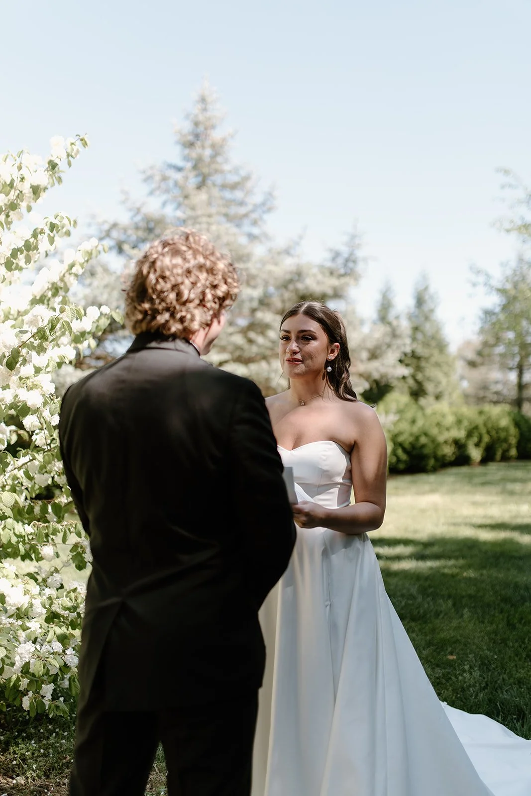 A bride and groom in a wedding ceremony outdoors, with the bride in a white strapless dress and the groom in a black suit, standing in front of white flowering bushes and trees under a clear blue sky.