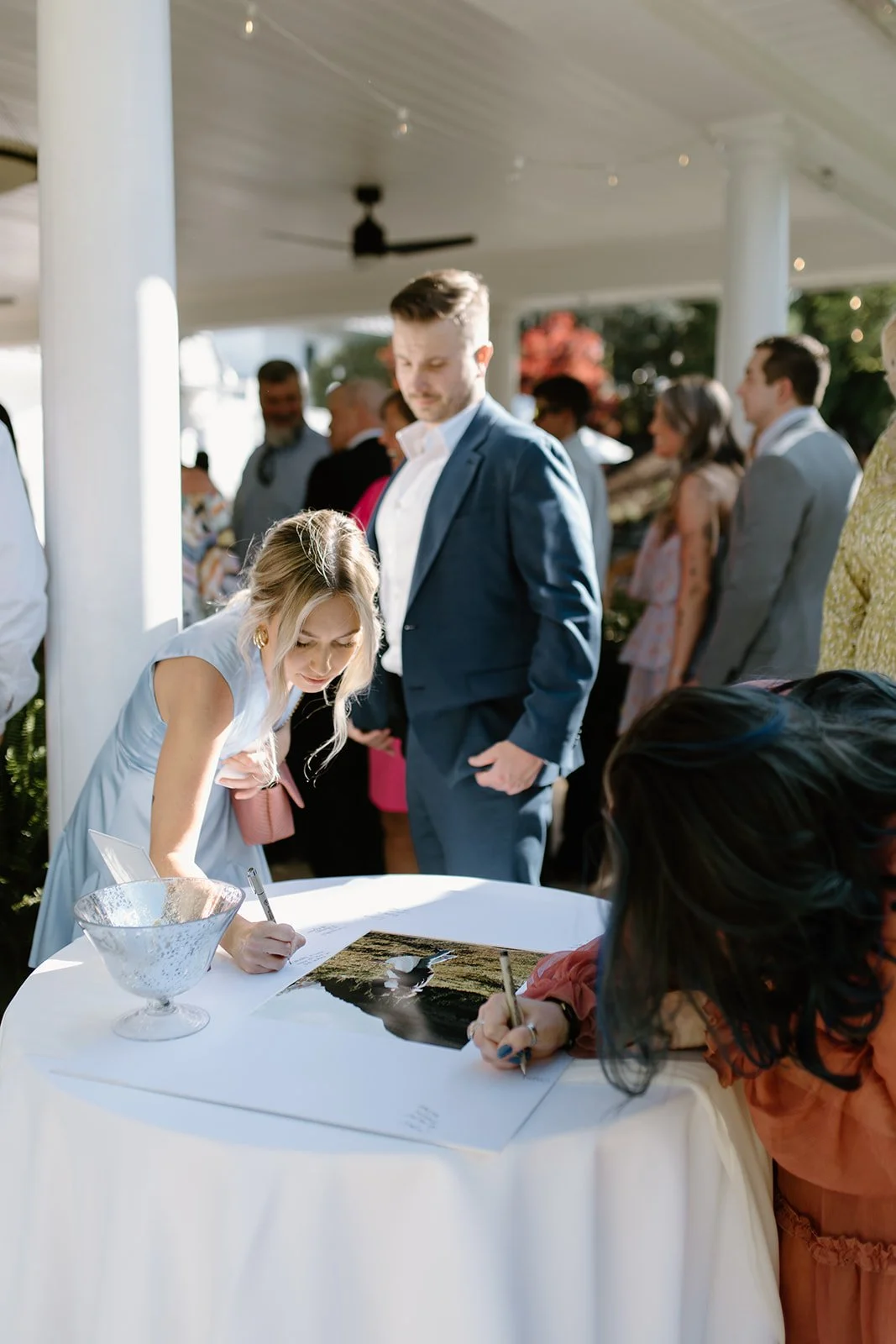 Two women signing guest books at a wedding reception, with guests mingling in the background under a covered outdoor area.