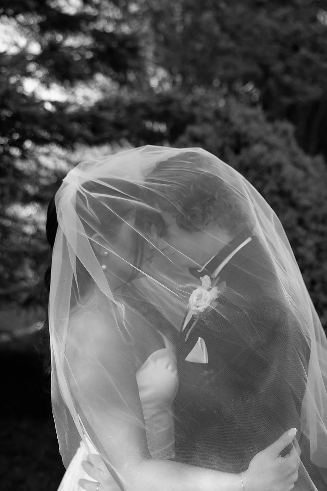 Black and white photo of a bride and groom embracing, with the bride wearing a veil that covers both their faces, outdoors with trees in the background.