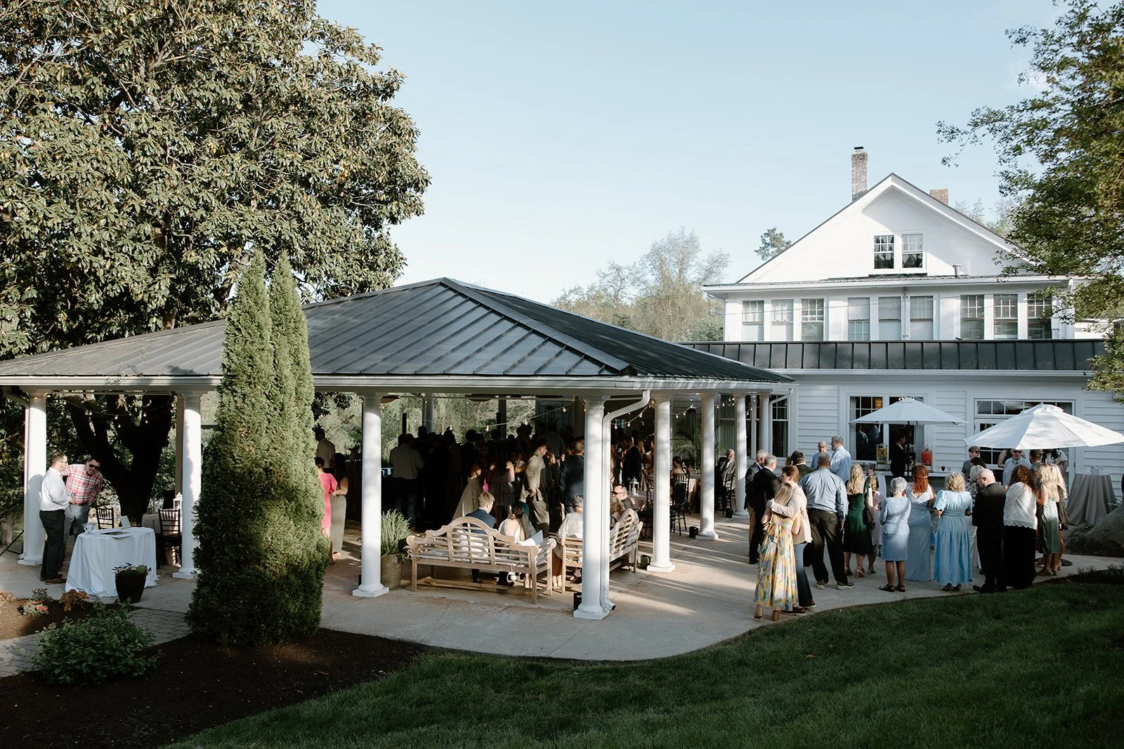 Outdoor wedding reception gathering under a pavilion, with guests socializing and seated, in front of a large white house with multiple windows and a black roof.