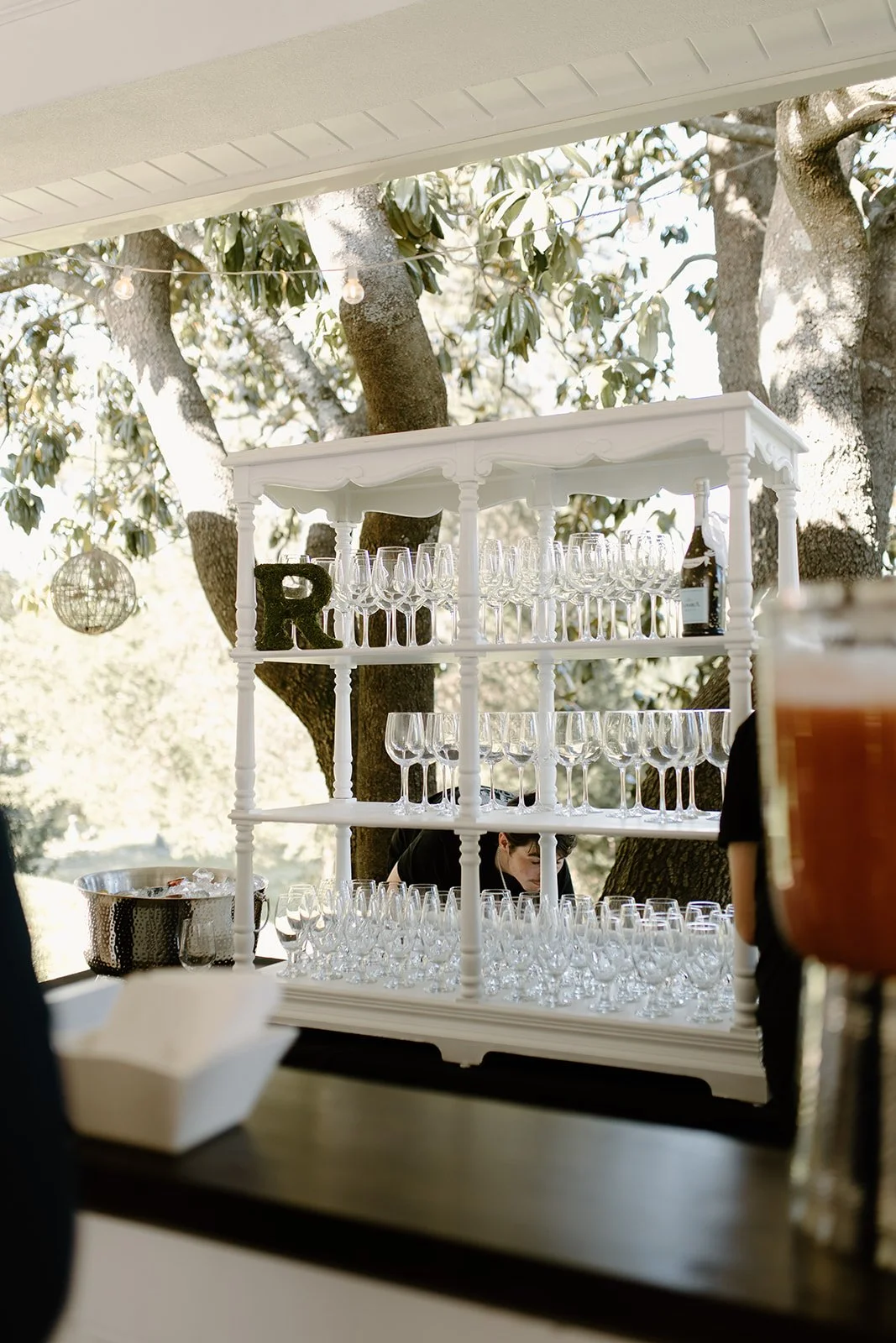 A white shelving unit with multiple rows of clean glassware, set against the background of trees and outdoor string lights, possibly at a wedding or outdoor event.