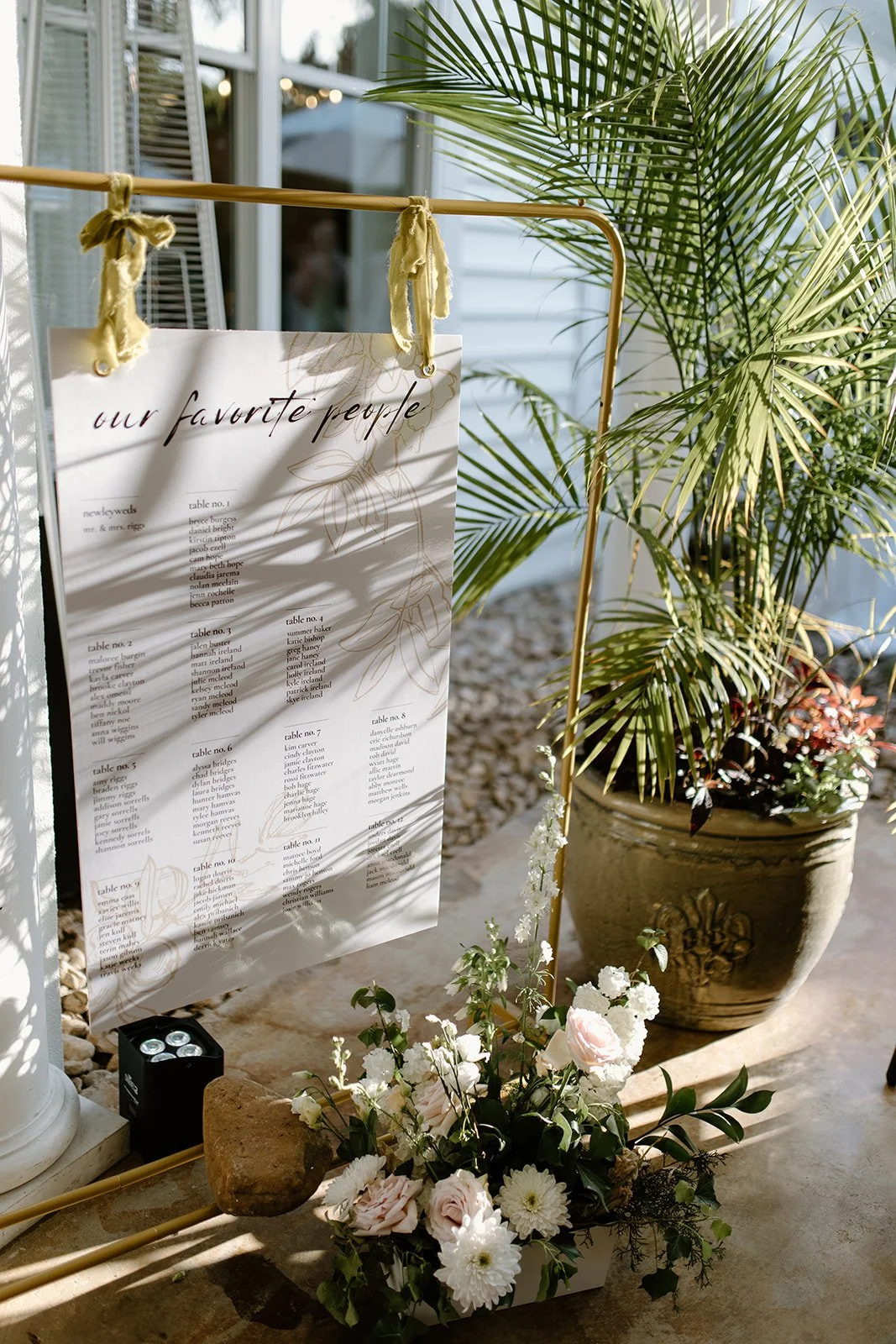 A wedding seating chart displayed on a large whiteboard with the title 'our favorite people,' surrounded by white flowers in a vase, a large potted plant, and decorative rocks, with sunlight casting shadows in a bright indoor space.