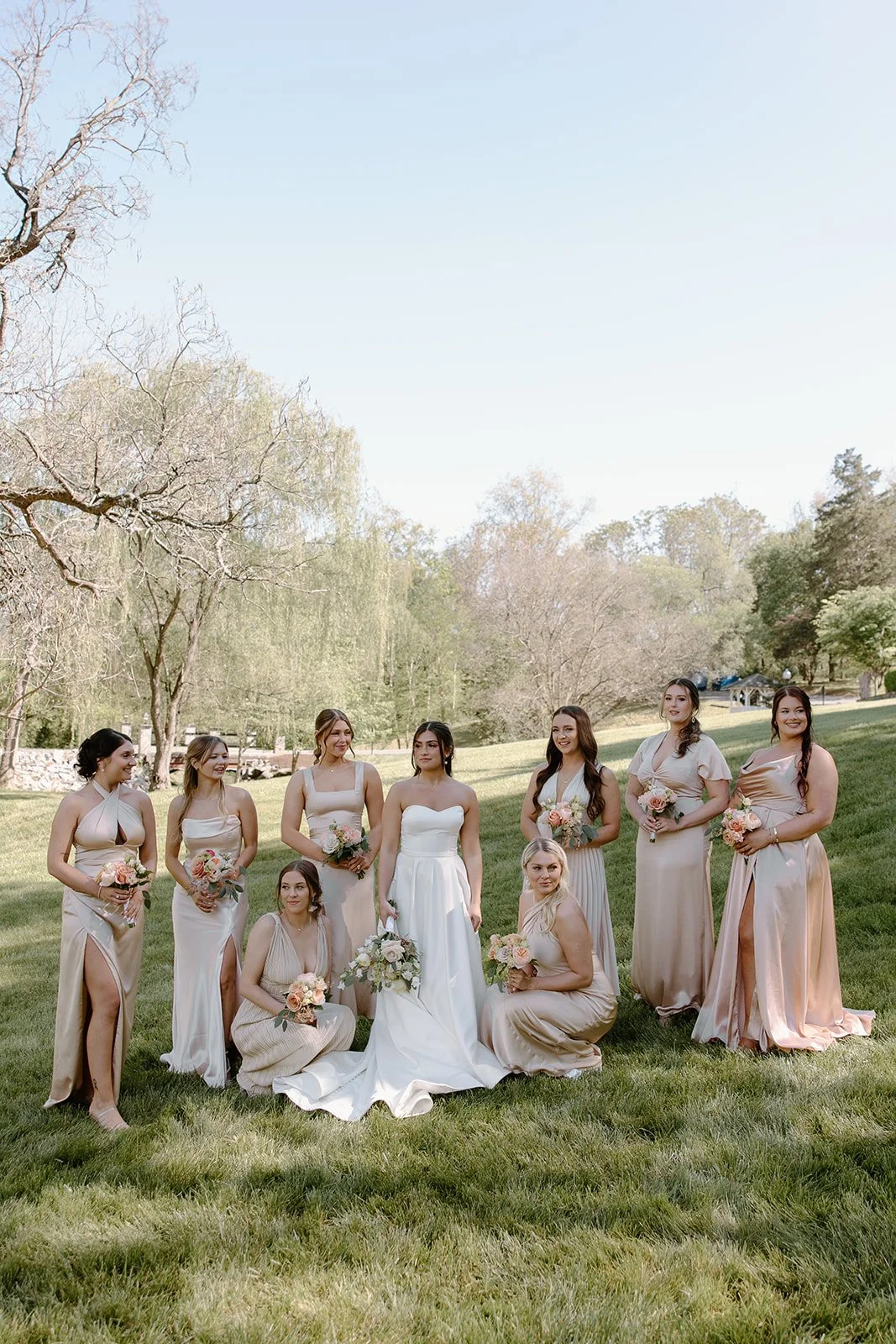 A bride in a white wedding dress surrounded by her bridesmaids in beige dresses holding bouquets, outdoors on a grassy area with trees in the background.