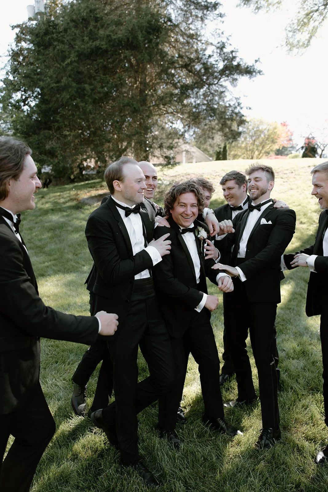 Group of men in tuxedos celebrating outdoors, smiling, and laughing on a grassy lawn with trees in the background.