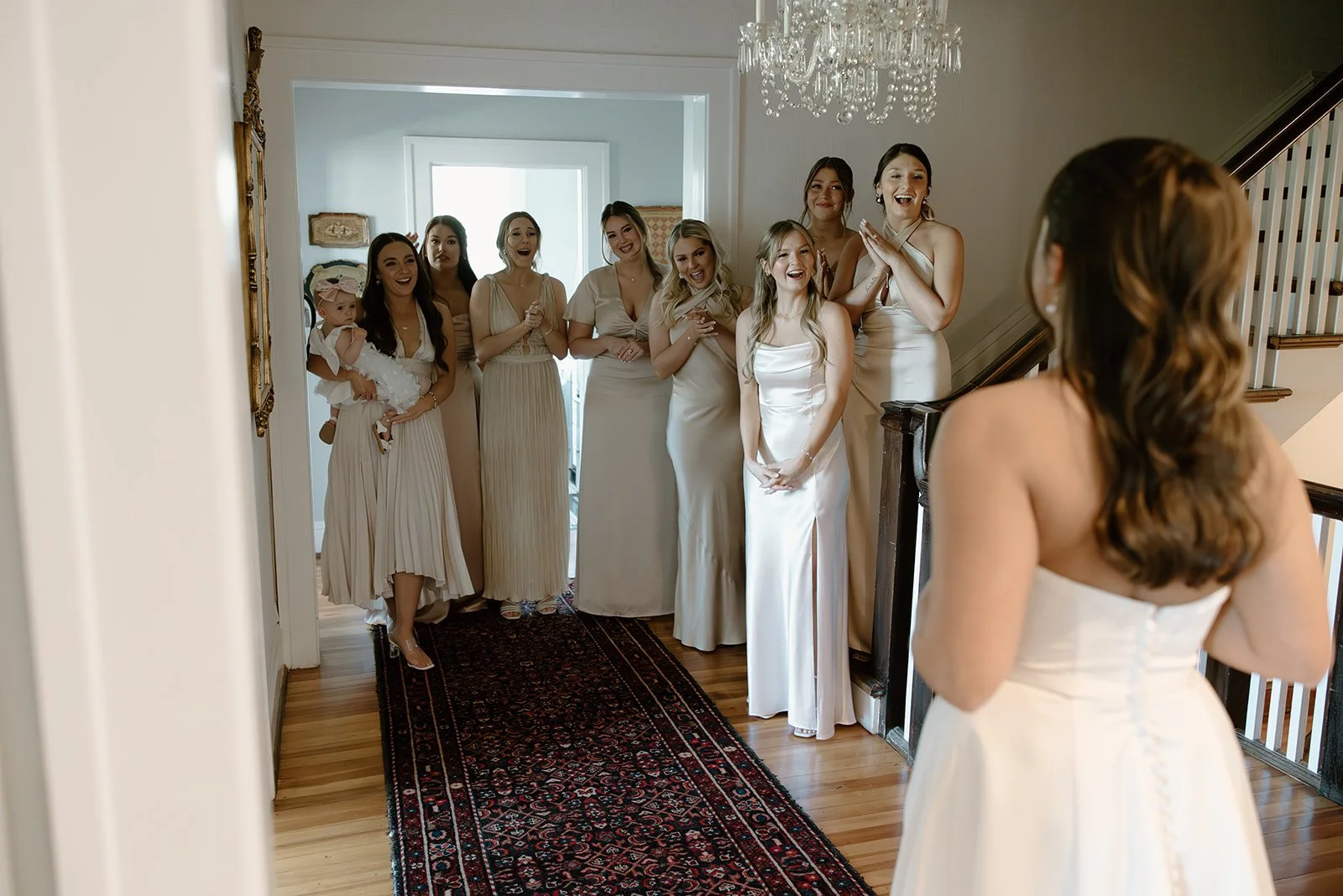 A bride in a white wedding dress standing in front of a group of women, likely bridesmaids, who are also dressed in white and reacting happily and excitedly.
