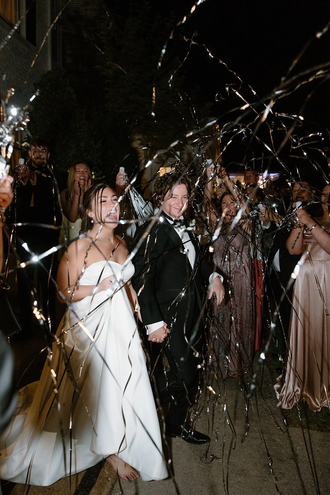 A bride and groom holding hands and smiling at night while surrounded by friends, celebrating with gold and silver streamers and lights.