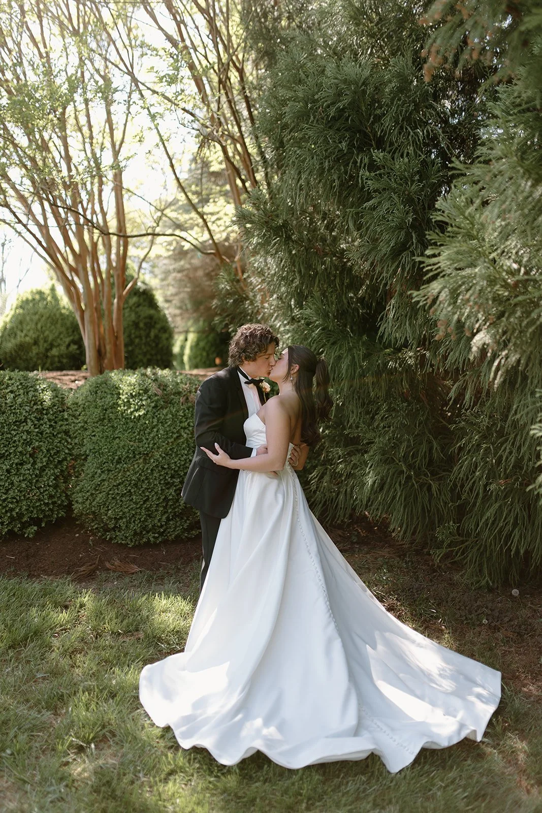 A newlywed couple kissing outdoors, surrounded by lush green trees and bushes, on a sunny day.