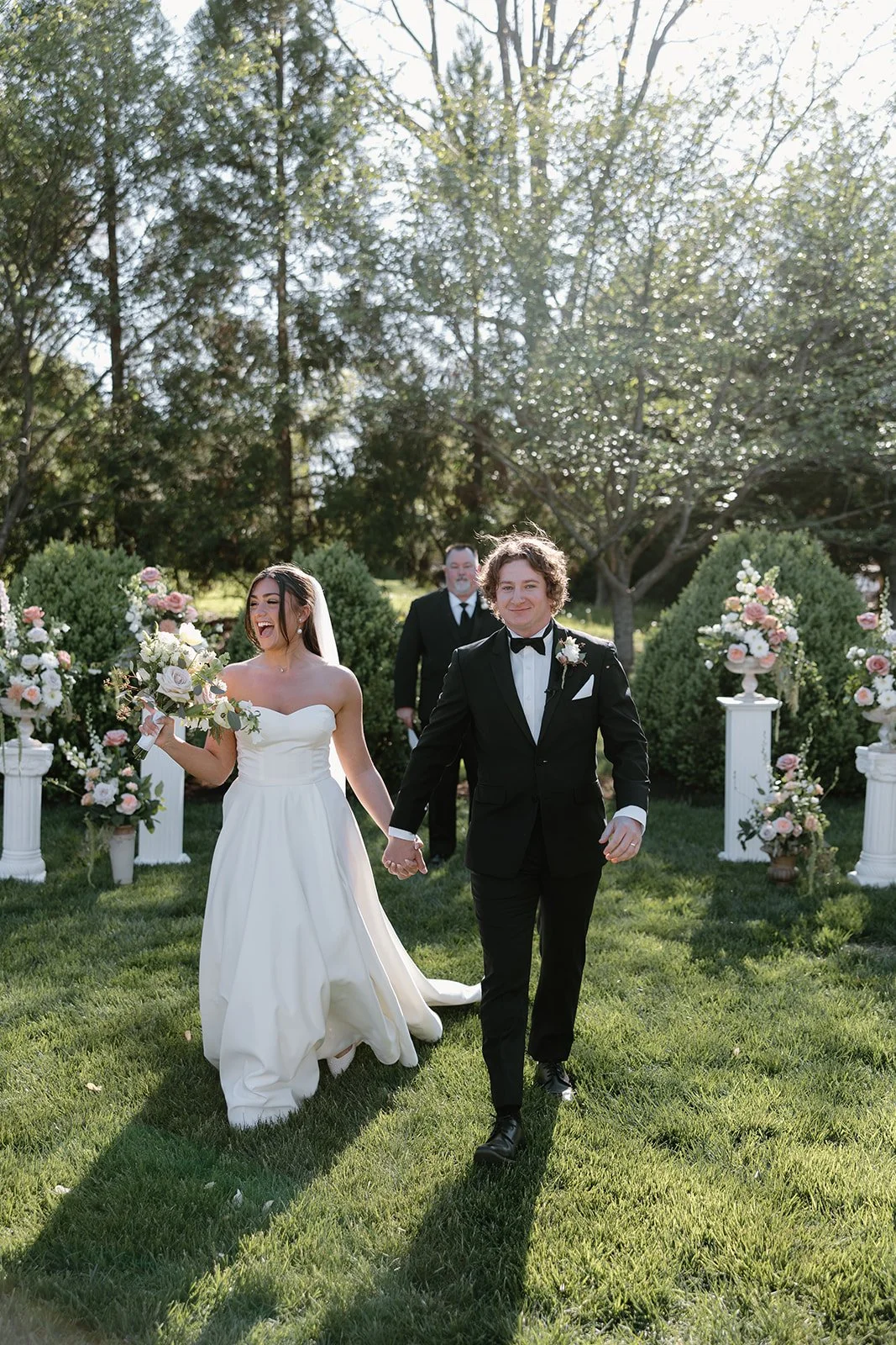 A newlywed couple walking hand-in-hand outdoors on a sunny day, with floral arrangements on pillars in the background, greenery, and trees.