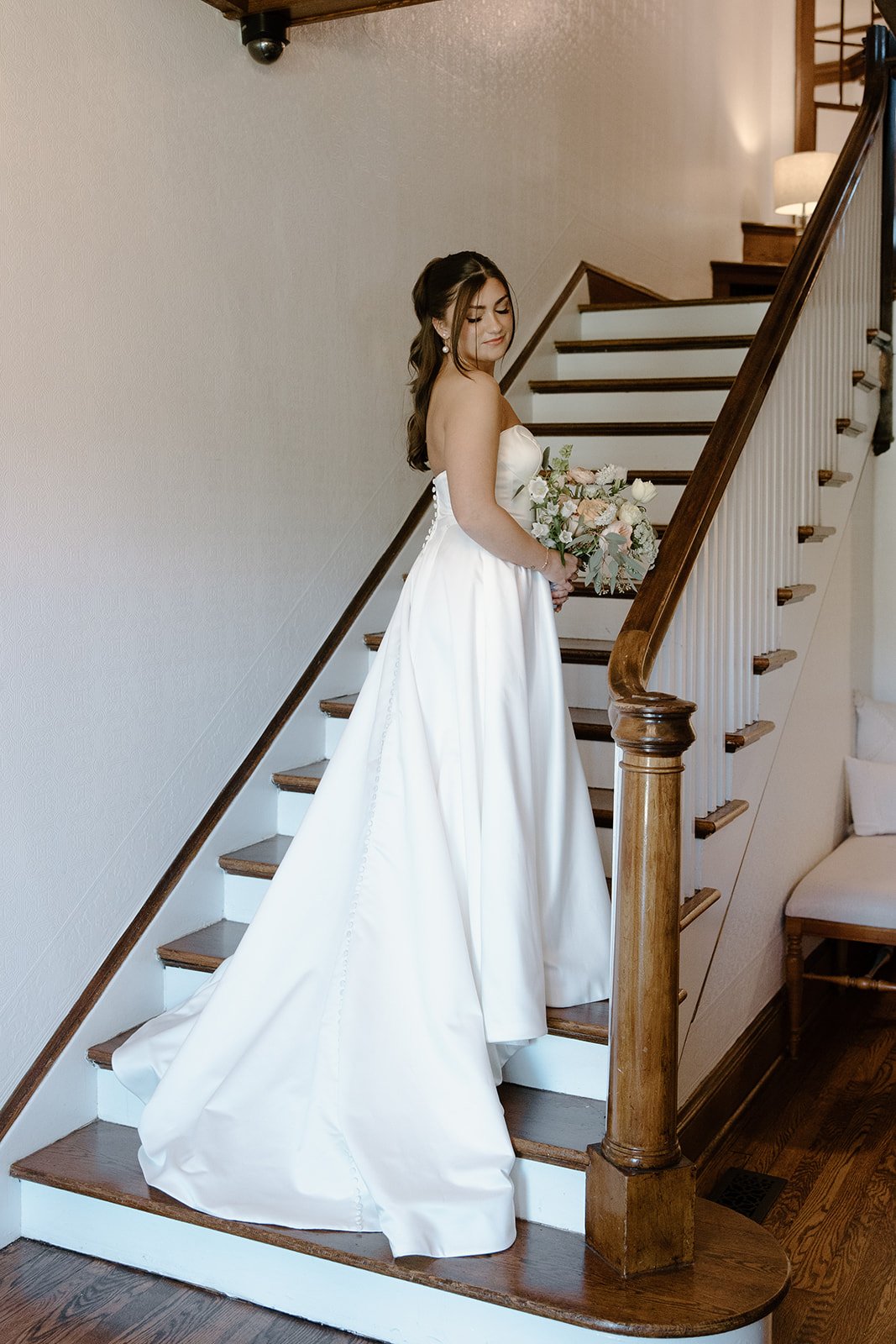 A bride in a white wedding gown holding a bouquet standing on staircase with wooden handrail in a home interior.
