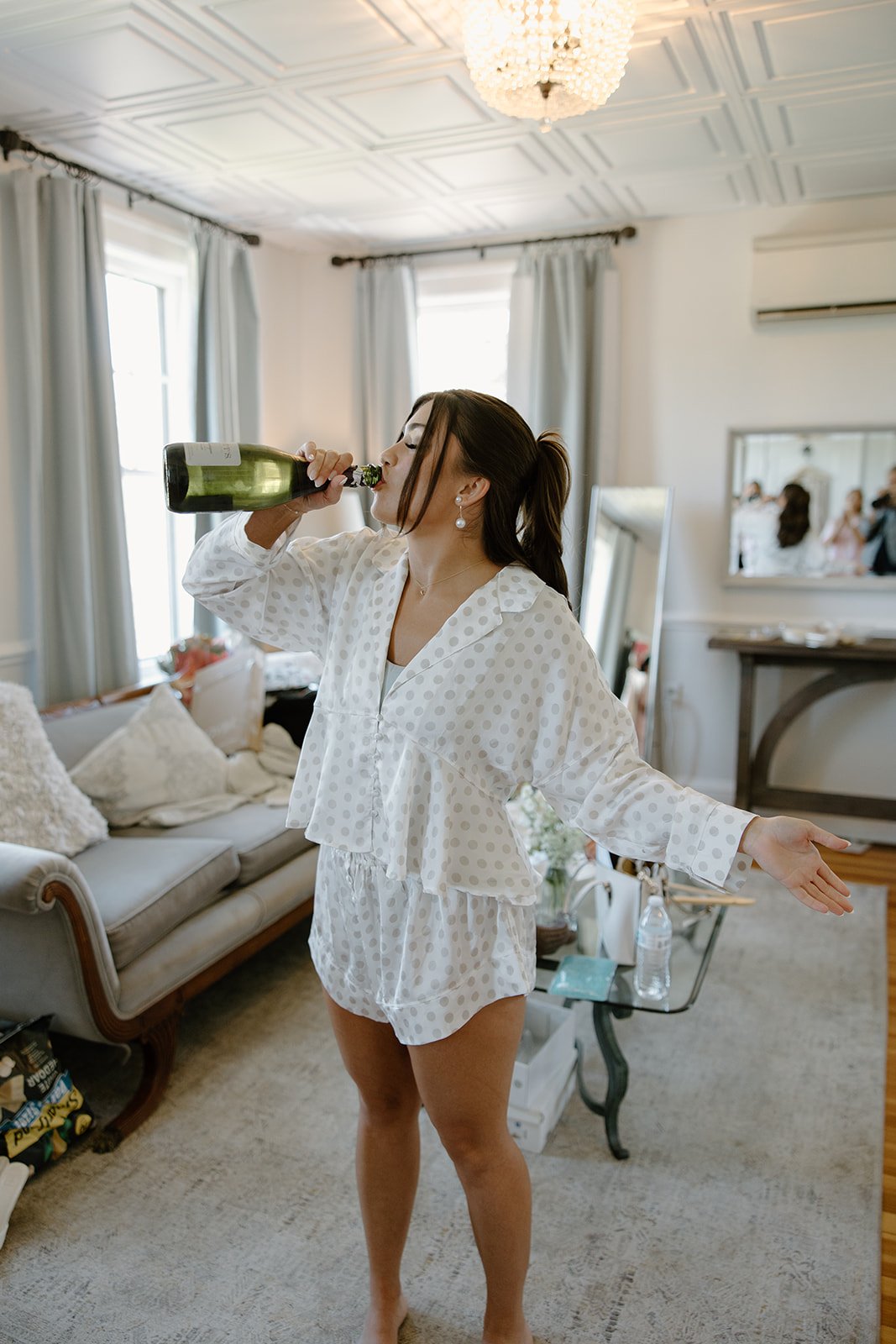 A woman in polka dot pajamas drinking from a bottle of champagne in a living room.
