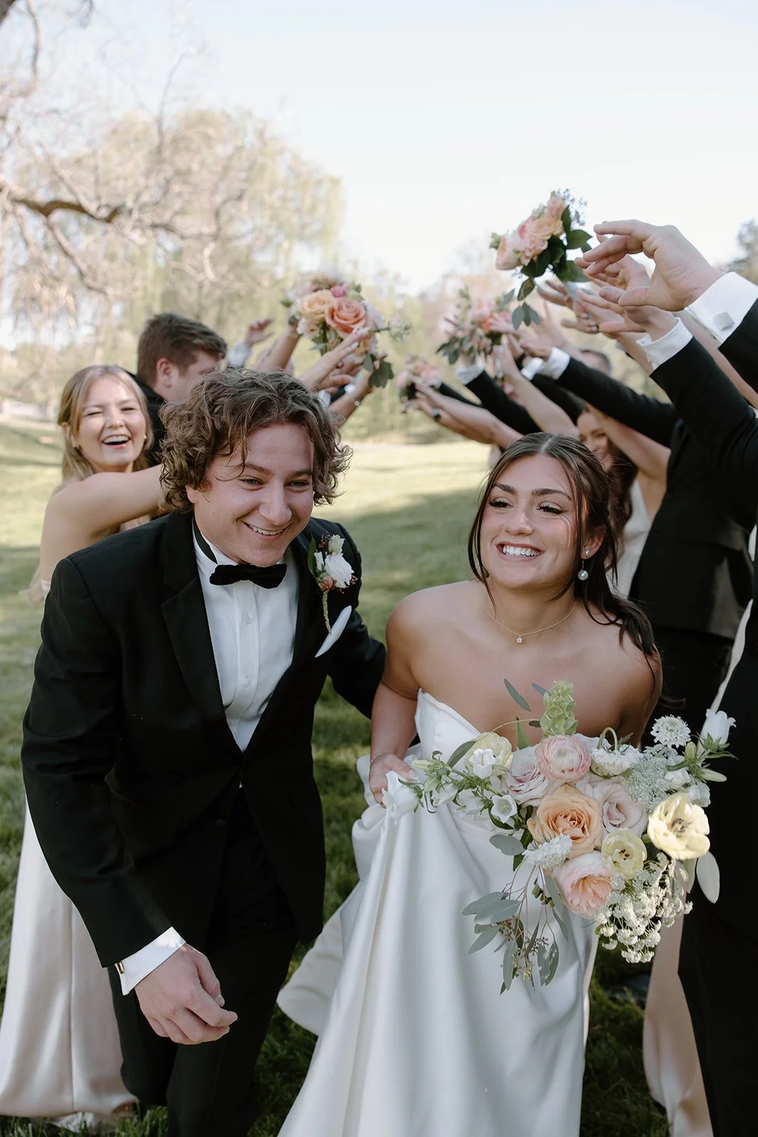 A bride and groom smiling and holding a bouquet, surrounded by friends and wedding party members holding pink and white flowers in an outdoor setting with trees and grass.