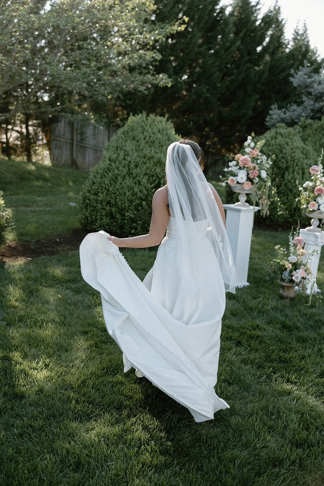 Bride in white wedding gown and veil outdoors with floral arrangements and greenery.