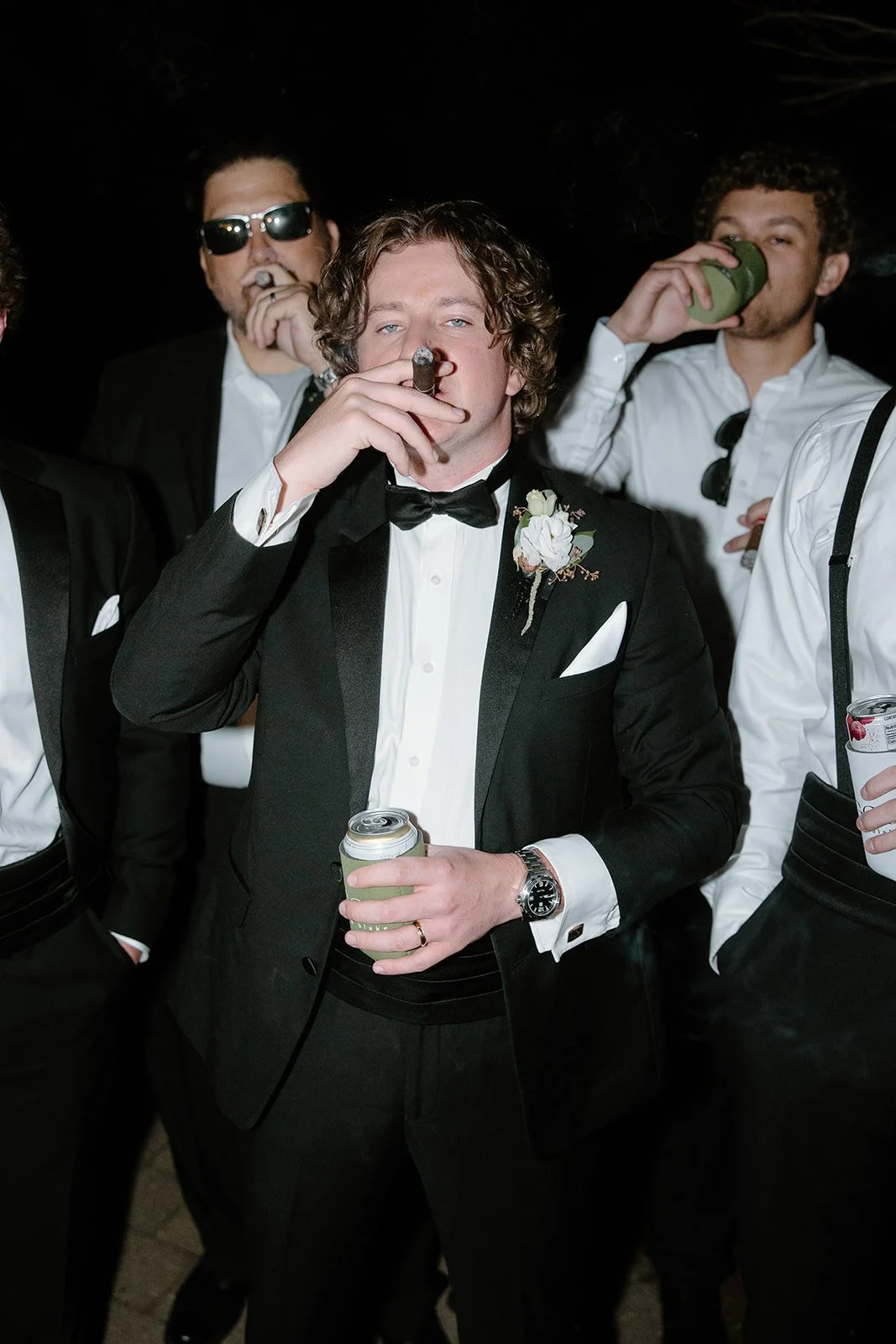 Group of men dressed in tuxedos, some smoking cigars and drinking, at a formal event.