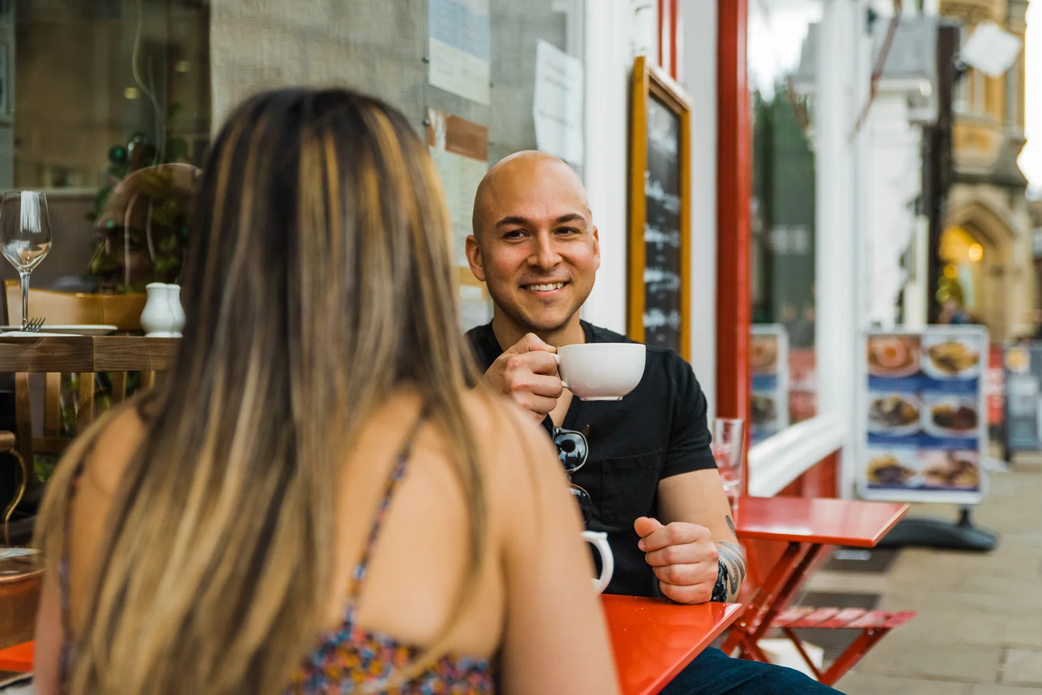 A man drinks coffee while smiling at his girlfriend during a couple photo session in Cambridge