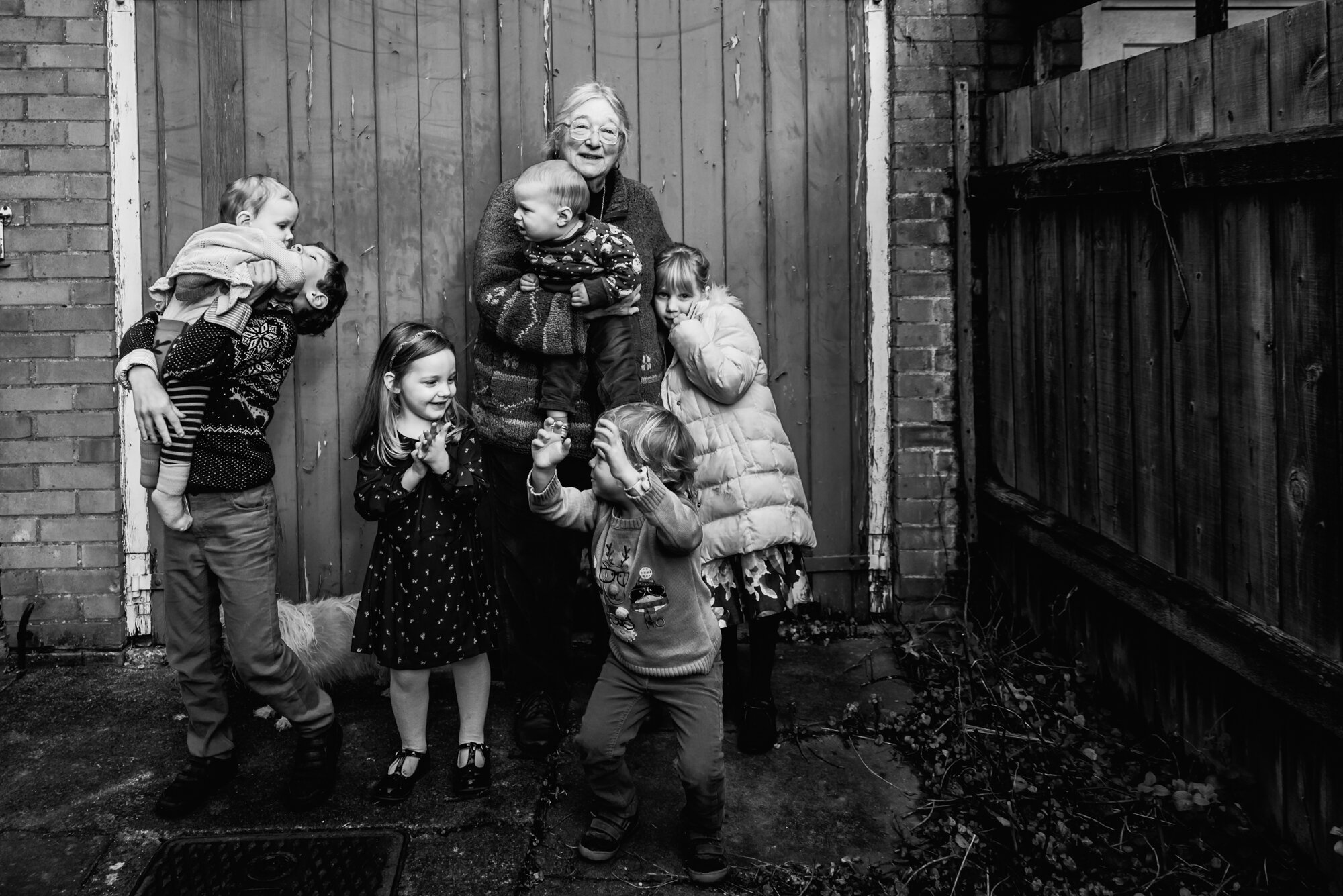  Black and white environmental portrait of a grandmother with all her grandchildren wearing Christmas jumpers taken in the garden of the family home in Cambridge. 