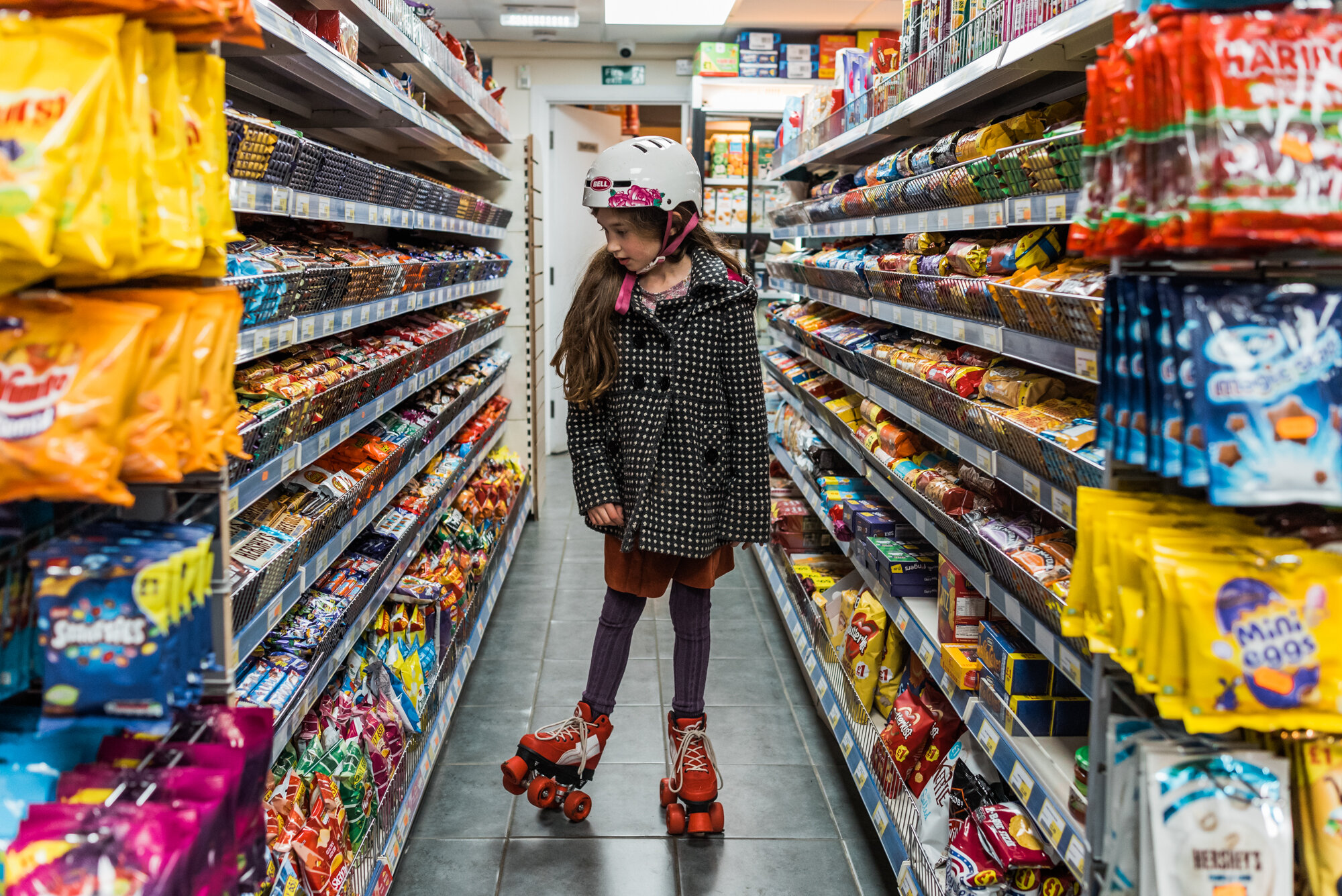  A young girl in pink roller skates and helmet stands in the aisle of a newsagent shop, looking at the rows of sweets and chocolates around her. 