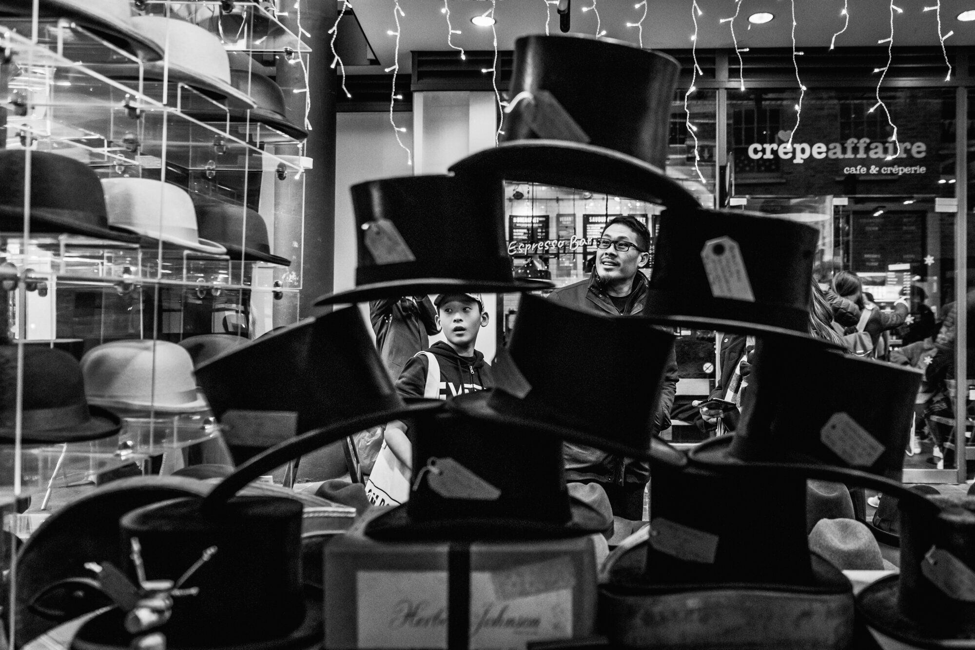  Black and white photograph of a Chinese man and his son is framed by a display of top hats at Spitalfields Market, London. 